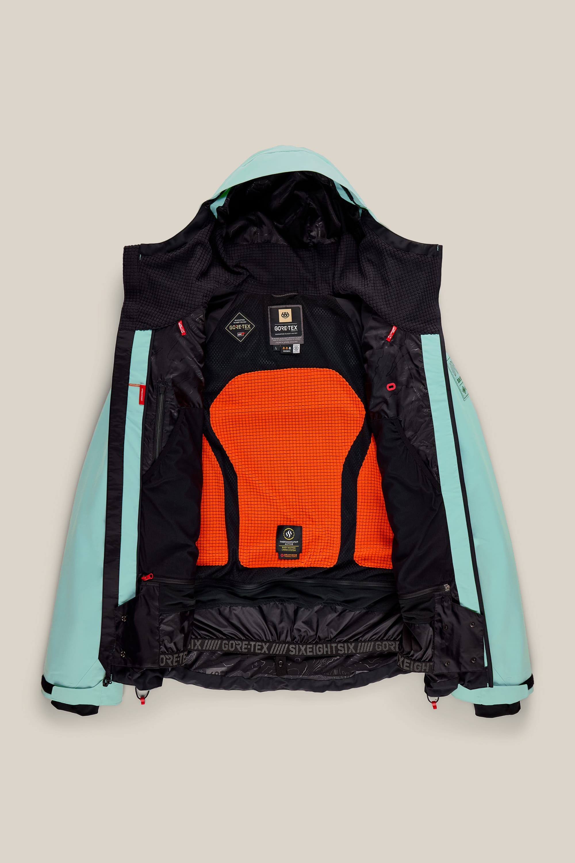 686 Men’s GORE-TEX® Dispatch™ Thermagraph® Jacket in light blue, featuring black and orange mesh lining, is displayed open to highlight the interior labels, pockets, and branding from 686 against a beige background.