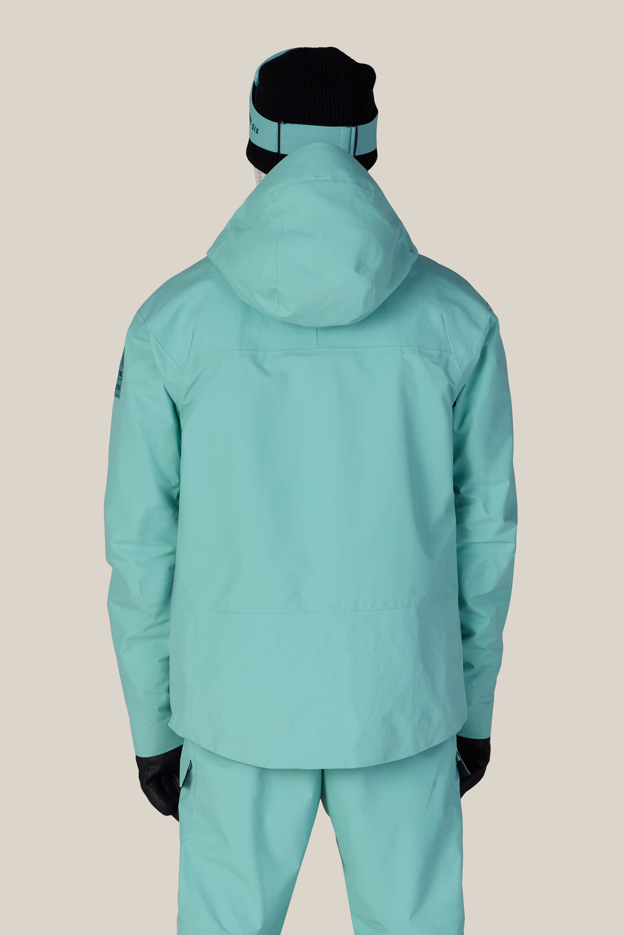 A person is shown from the back against a beige background, wearing the 686 Men’s GORE-TEX® Dispatch™ Thermagraph® Jacket in light blue with matching pants, black gloves, and a black beanie with a light blue band.