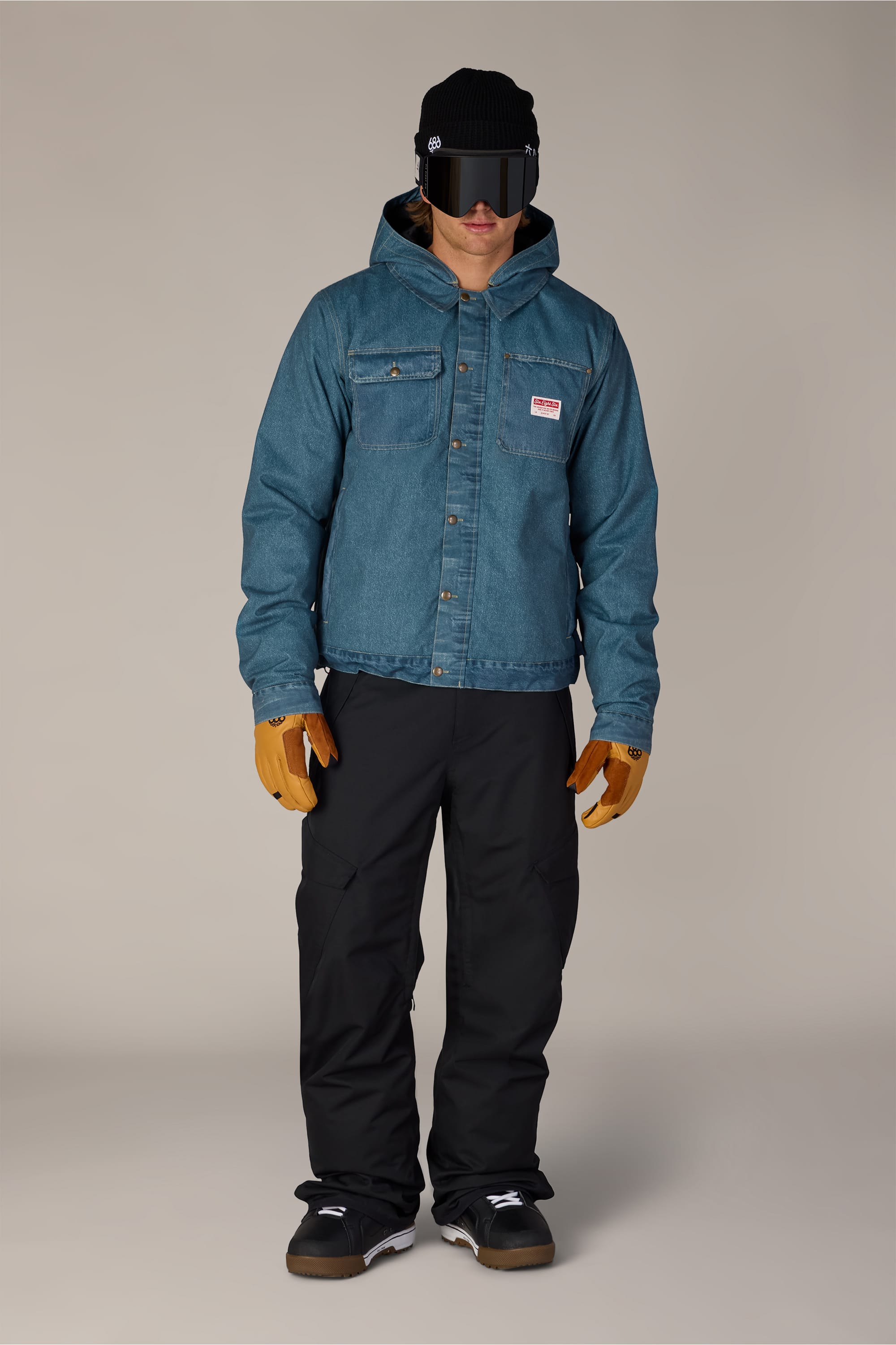 A person wearing the 686 Men’s Deconstructed™ Denim Insulated Jacket in teal, paired with black pants, brown gloves, black shoes, a black visor, and a black beanie stands against a plain background.