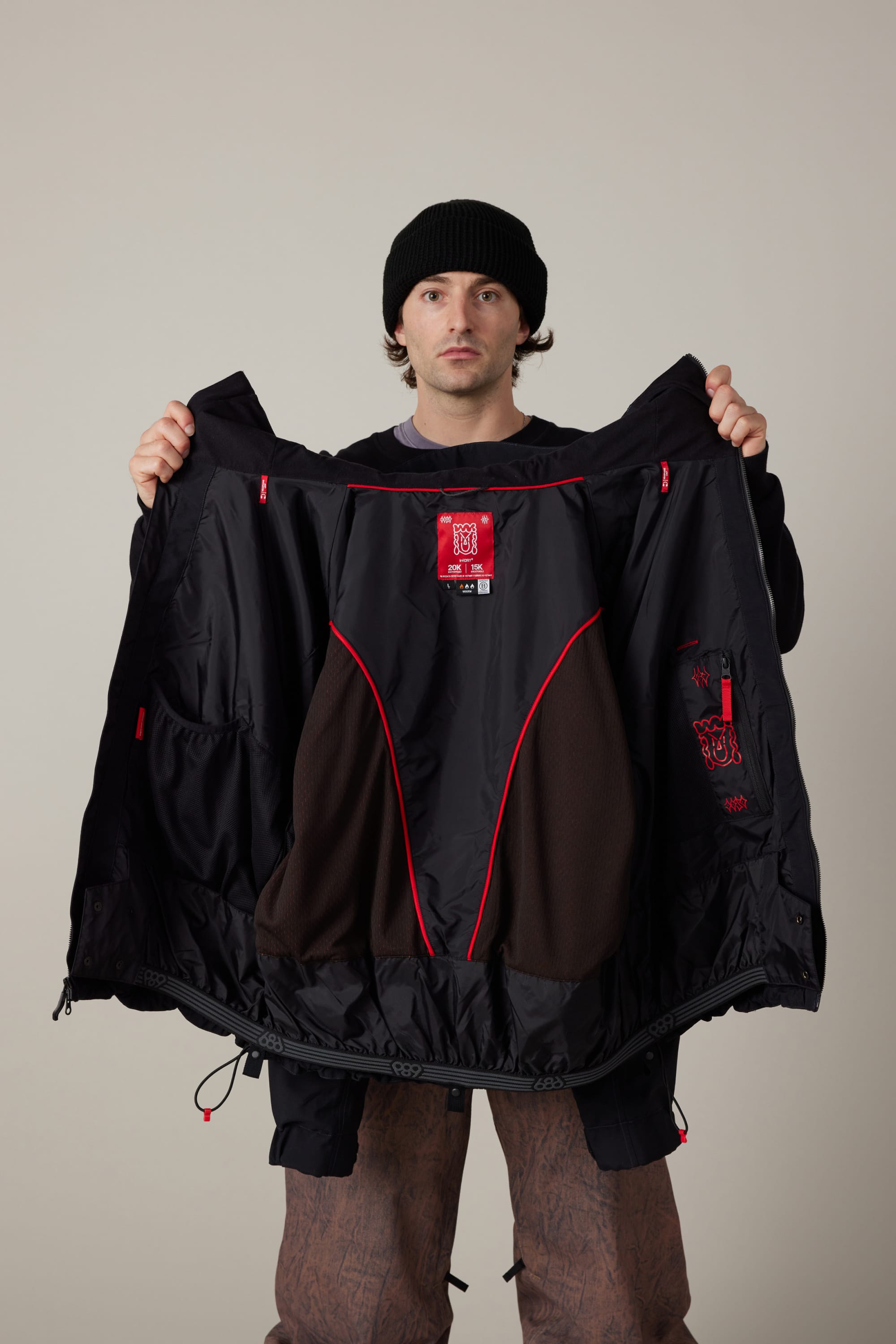 A person in a black beanie and patterned pants holds open the 686 Men’s 686 Dojo® Shell Jacket by 686, displaying its interior lining and red trim against a plain background.