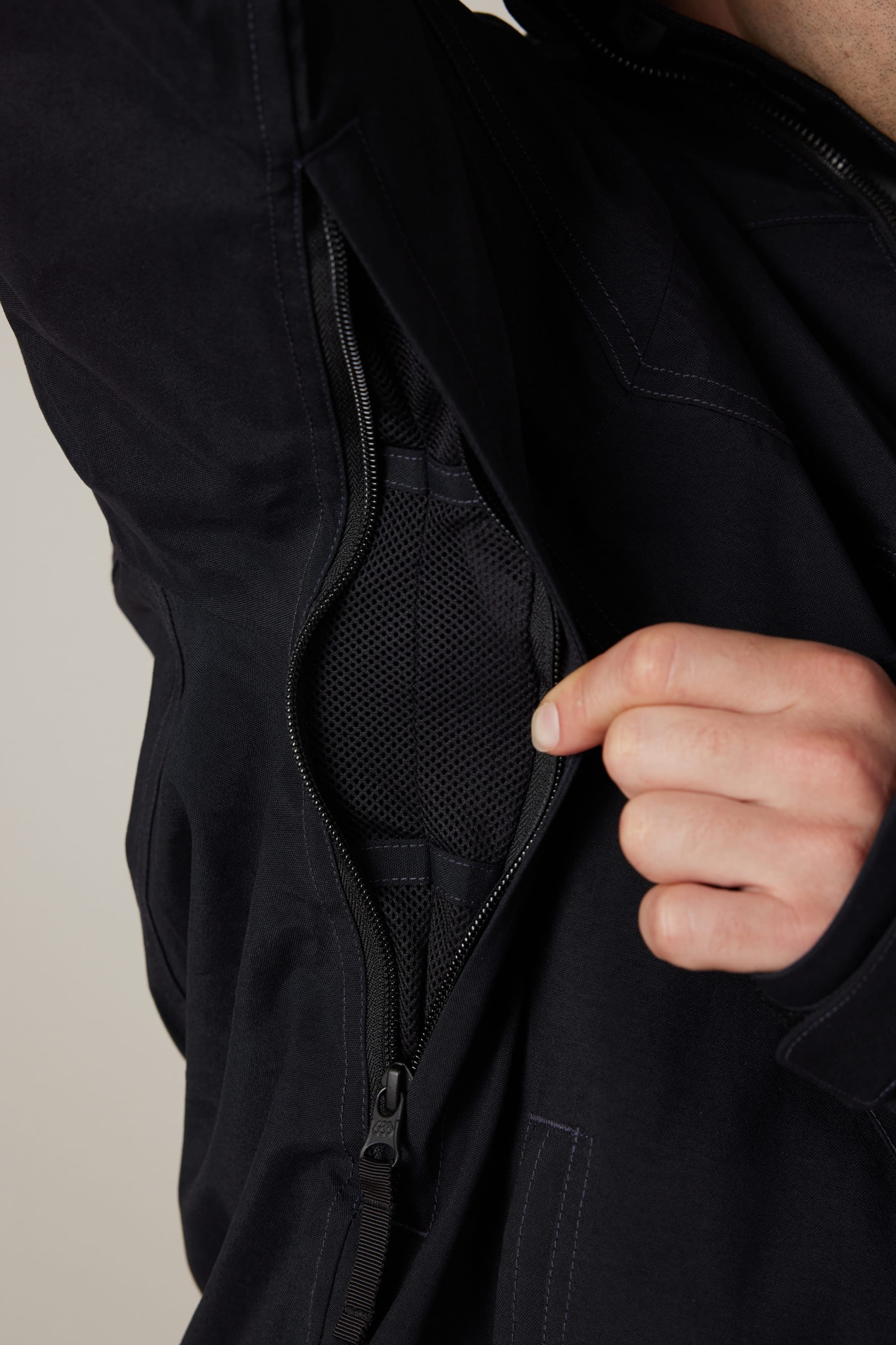 A person unzips the 686 Men’s 686 Dojo® Shell Jacket in black, revealing black mesh fabric lining inside. Only their hand and part of the jacket are visible.