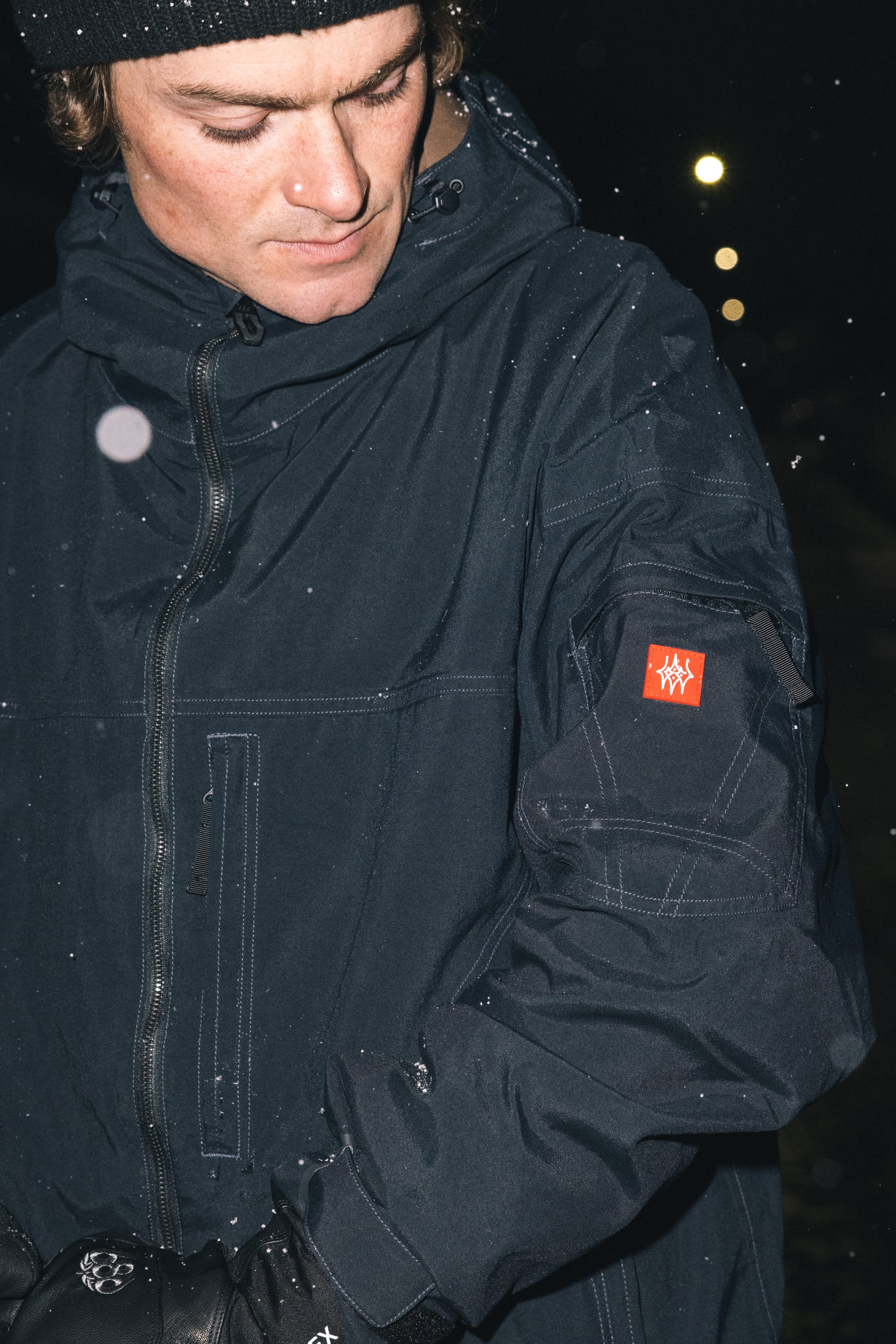 A person in the 686 Men’s 686 Dojo® Shell Jacket and a black beanie looks down at their sleeve, which features a red patch with a white symbol. Snowflakes fall around them, and it is nighttime.