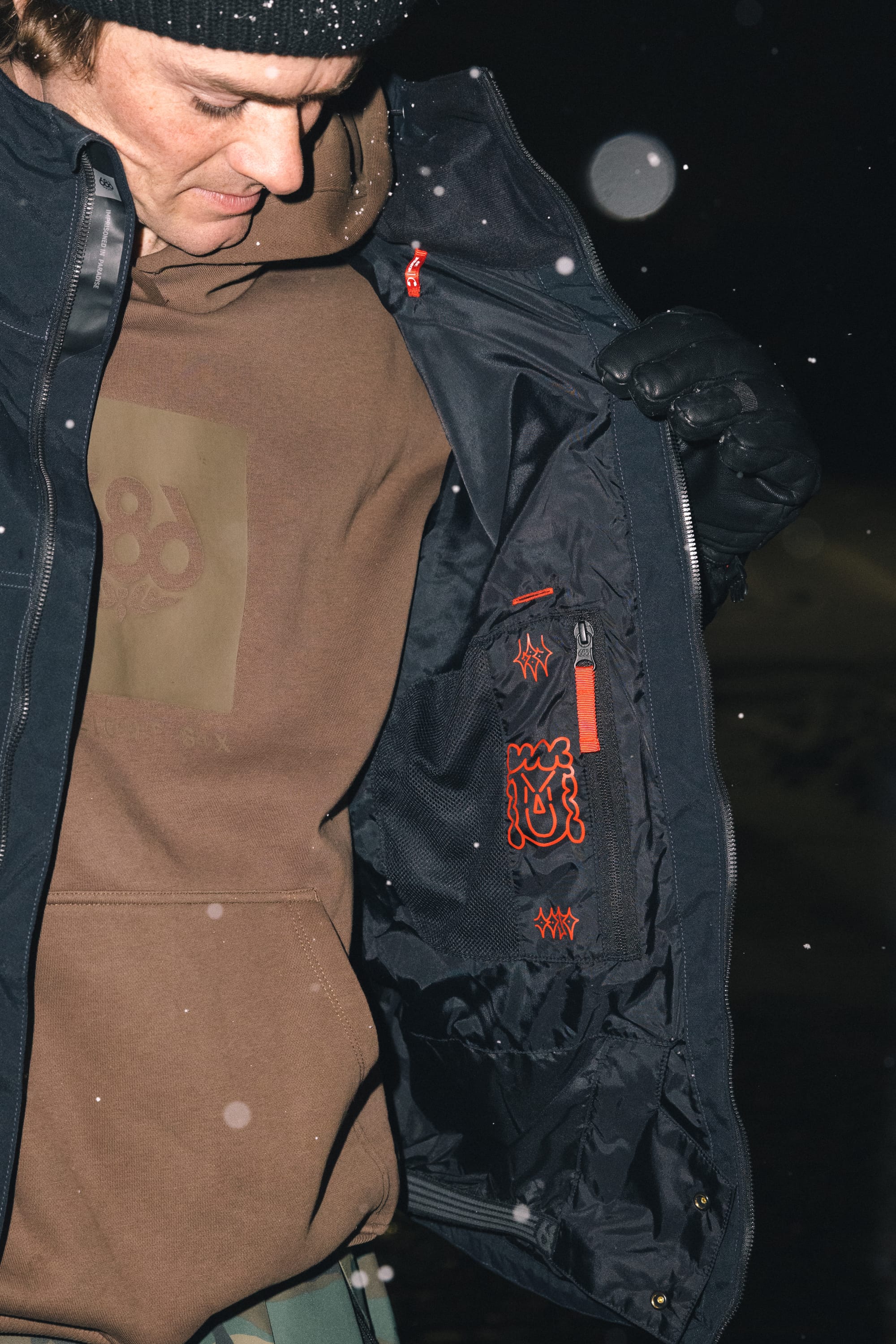 Wearing winter gear, a person opens their black 686 Men’s 686 Dojo® Shell Jacket to reveal inner pockets with red designs as snow falls, looking down at the inside of the jacket.