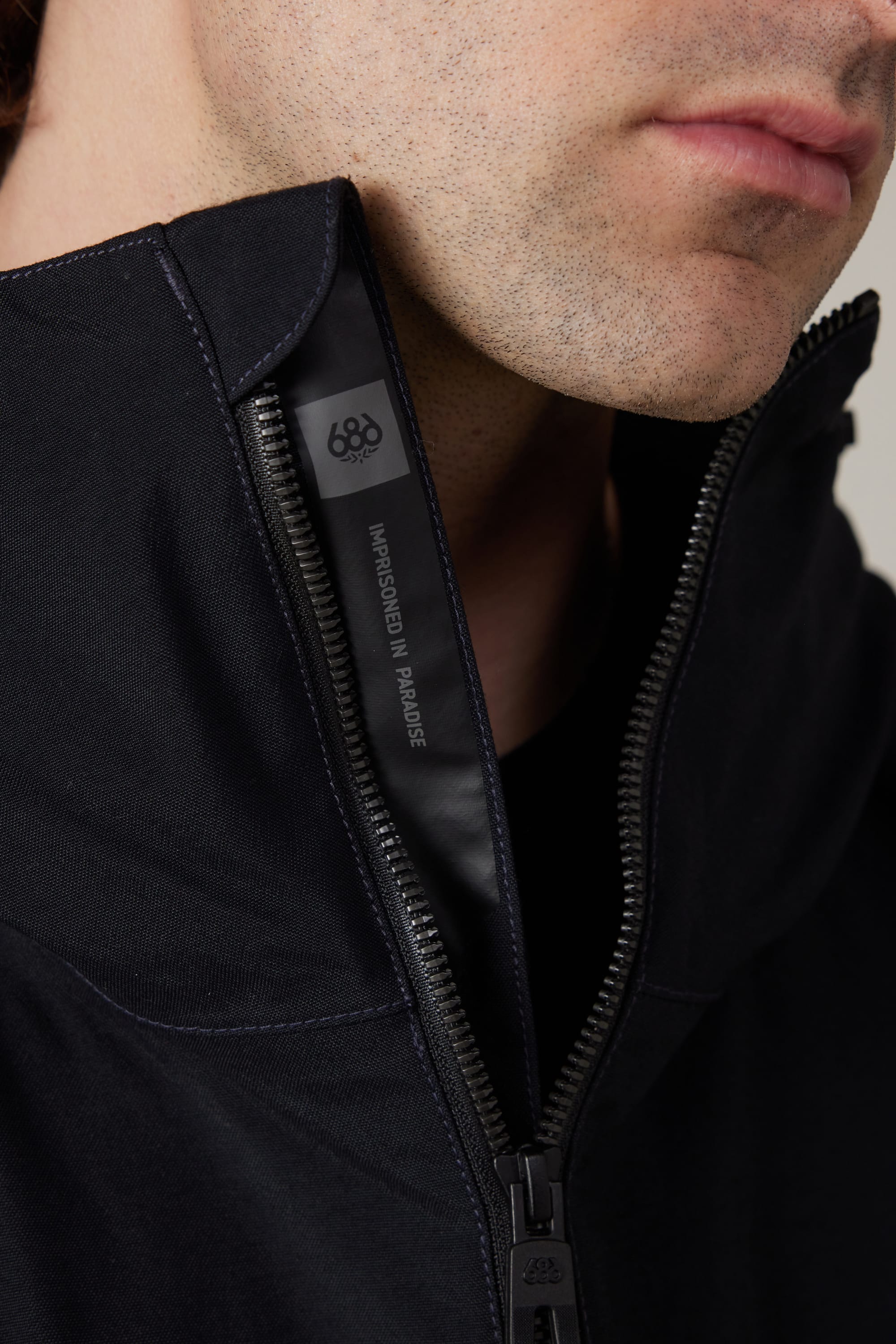Close-up of a person wearing a partially unzipped 686 Men’s 686 Dojo® Shell Jacket in black, revealing a tag inside the collar that reads IMAGINED IN PARADISE and the number 686. Only the lower half of the person’s face is visible.