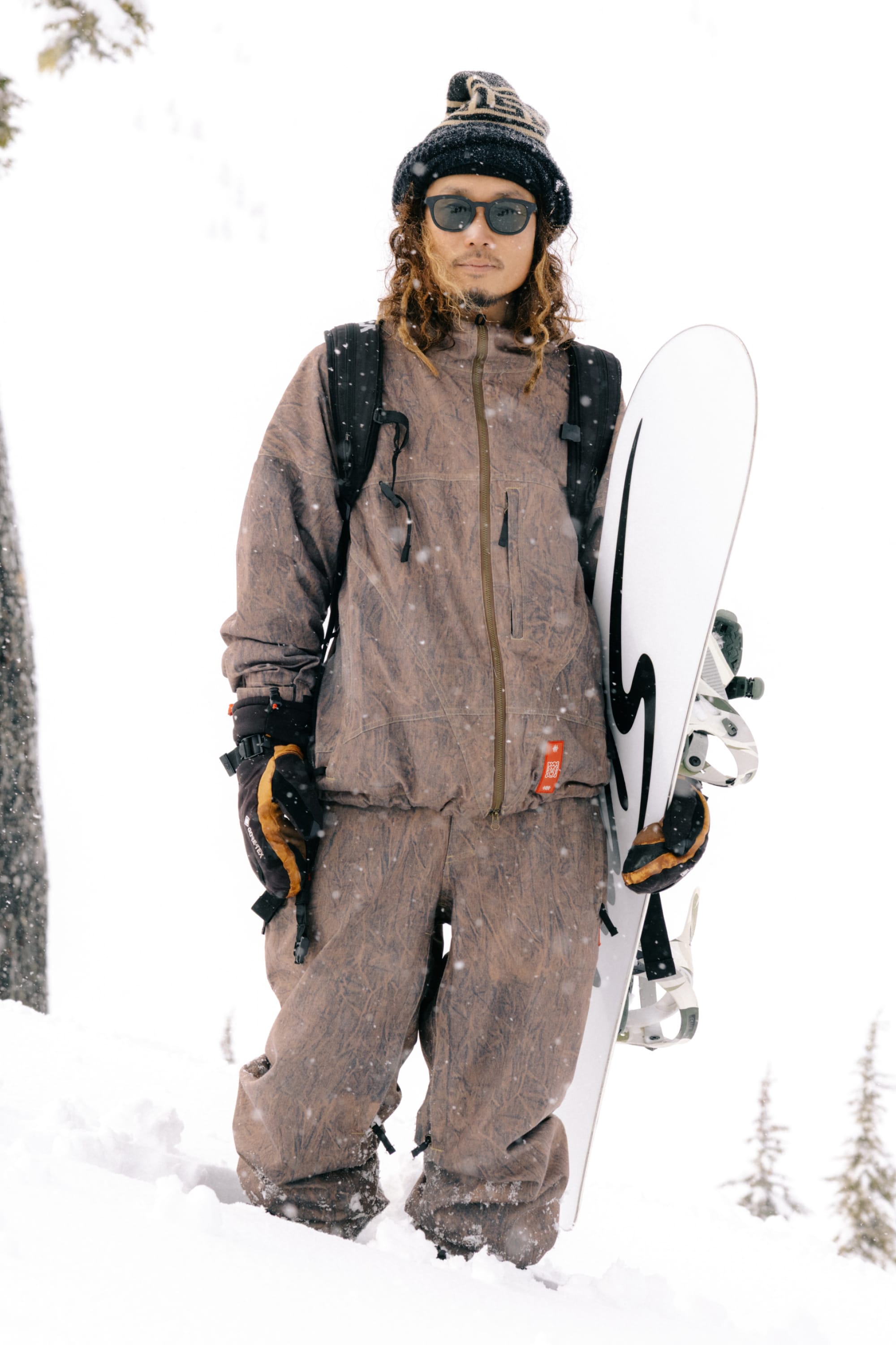 A person in brown snow gear, including the 686 Men’s 686 Dojo® Shell Jacket by 686, gloves, sunglasses, and a beanie, stands in the snow holding a snowboard, with trees and falling snow in the background.