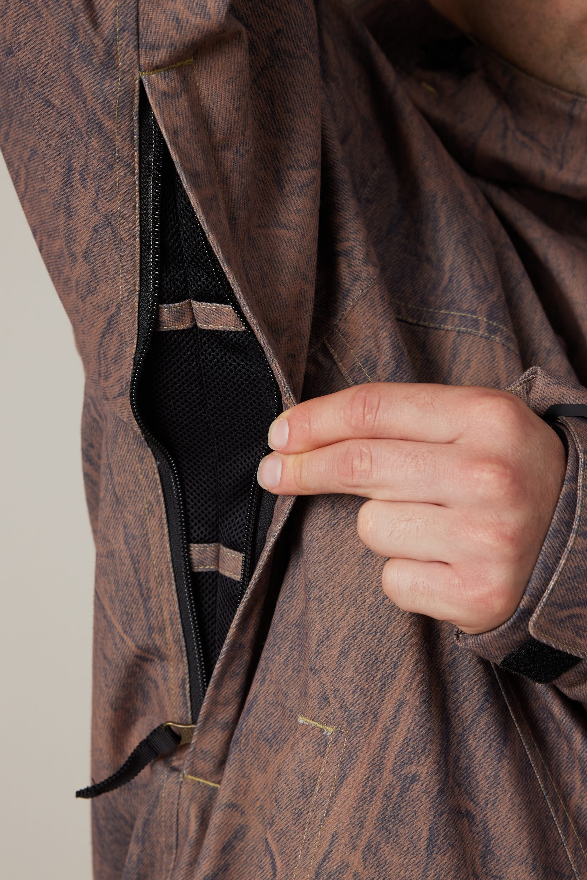 A person pulls open a zippered vent under the arm of a brown, patterned 686 Men’s Dojo® Shell Jacket, revealing a black mesh lining inside.