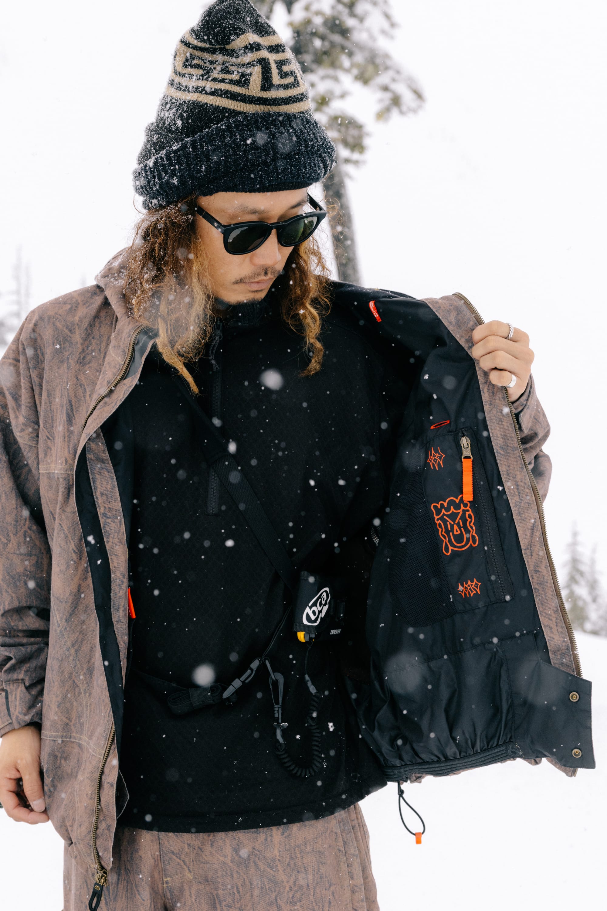 A person wearing sunglasses, a patterned beanie, and the 686 Men’s 686 Dojo® Shell Jacket by 686 stands in the snow, holding their jacket open to reveal the inner lining and multiple pockets as snow falls around them.