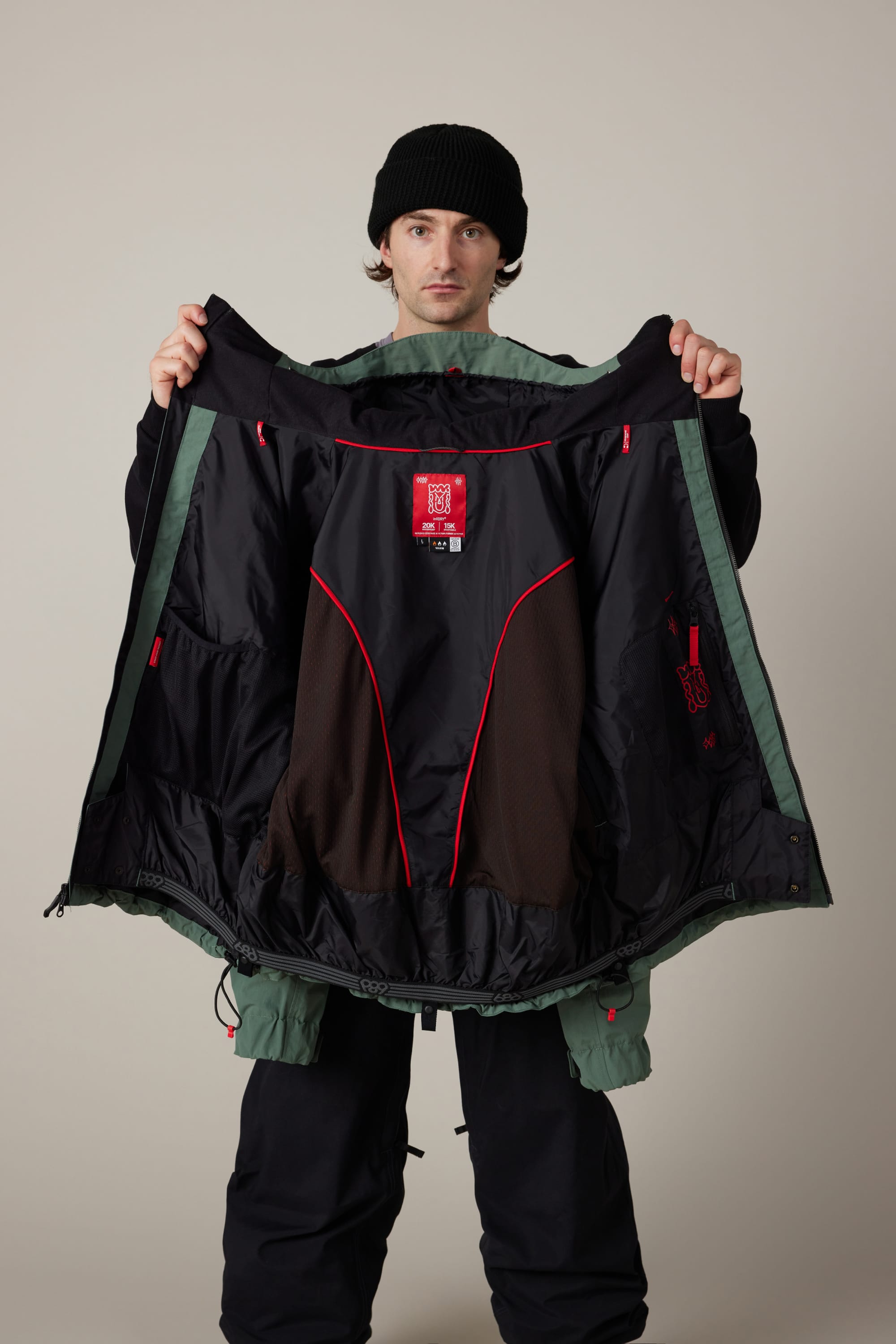 A person in a black beanie displays the 686 Men’s 686 Dojo® Shell Jacket by holding it open to reveal its black and red-lined interior, standing against a plain light background.