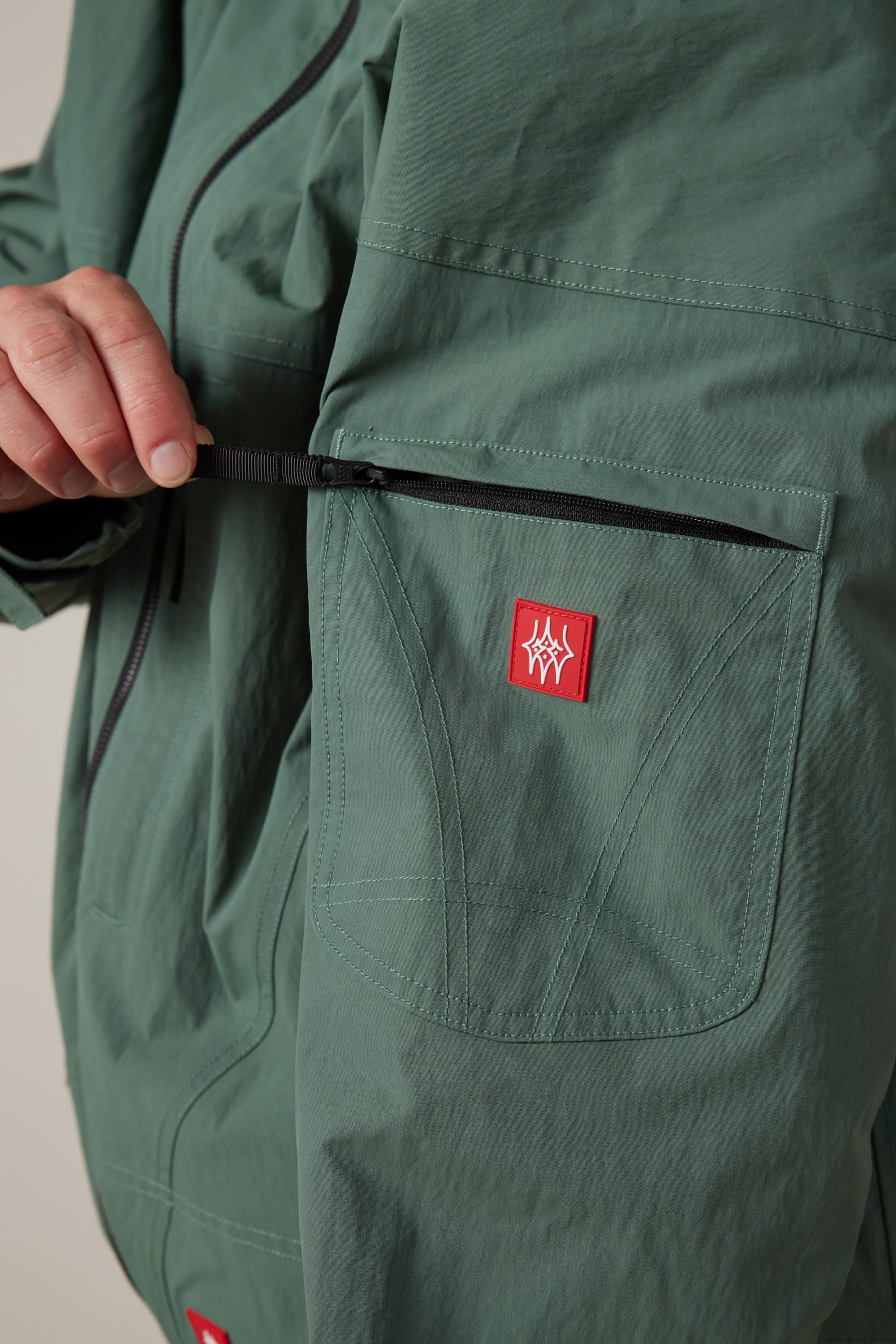 A person opens the zippered pocket of a green 686 Men’s 686 Dojo® Shell Jacket, which features a red rectangular patch with a white abstract logo.