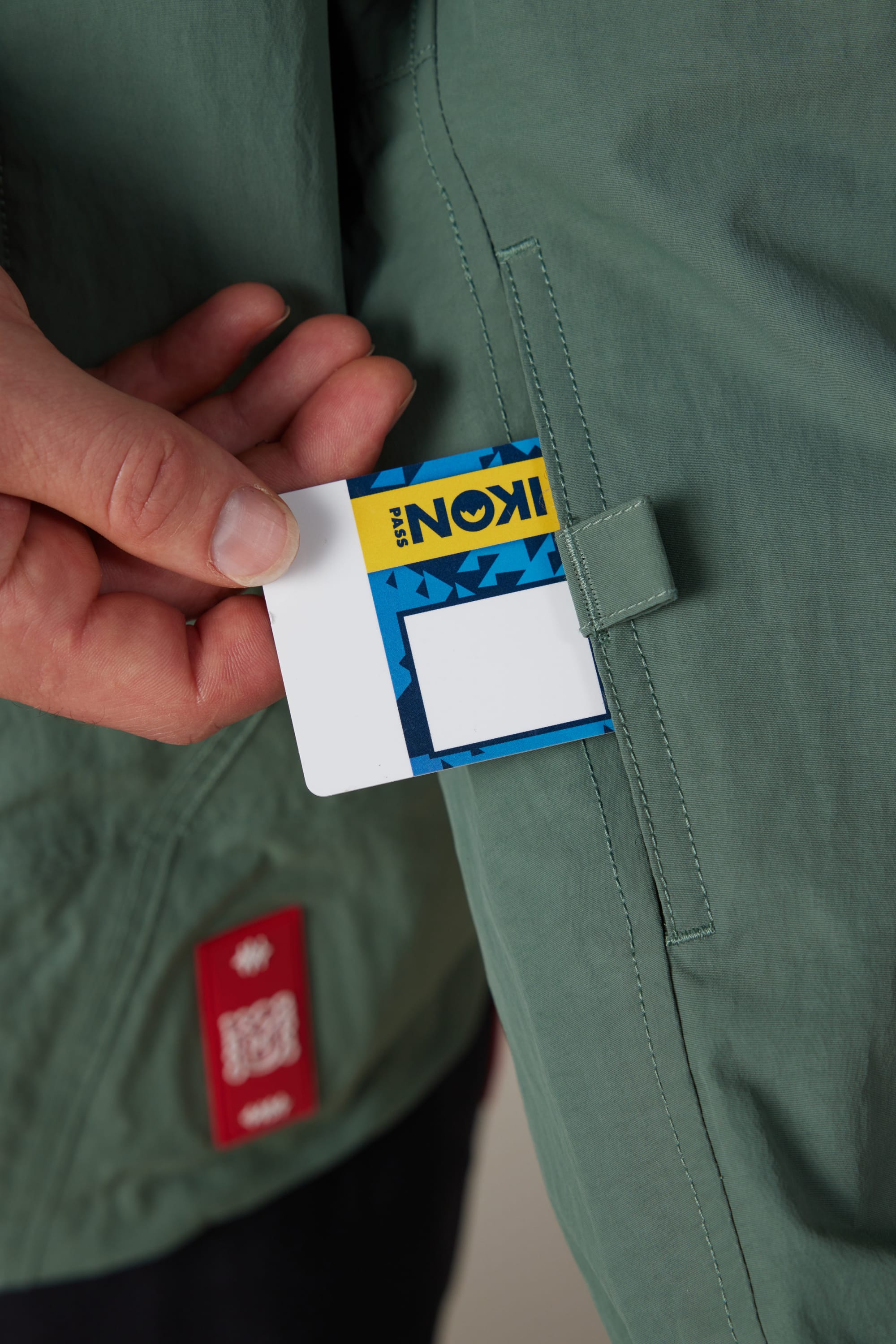 A hand holds a rectangular card with colorful patterns and the word IKON near the pocket of a men’s 686 Dojo® Shell Jacket by 686 in green.