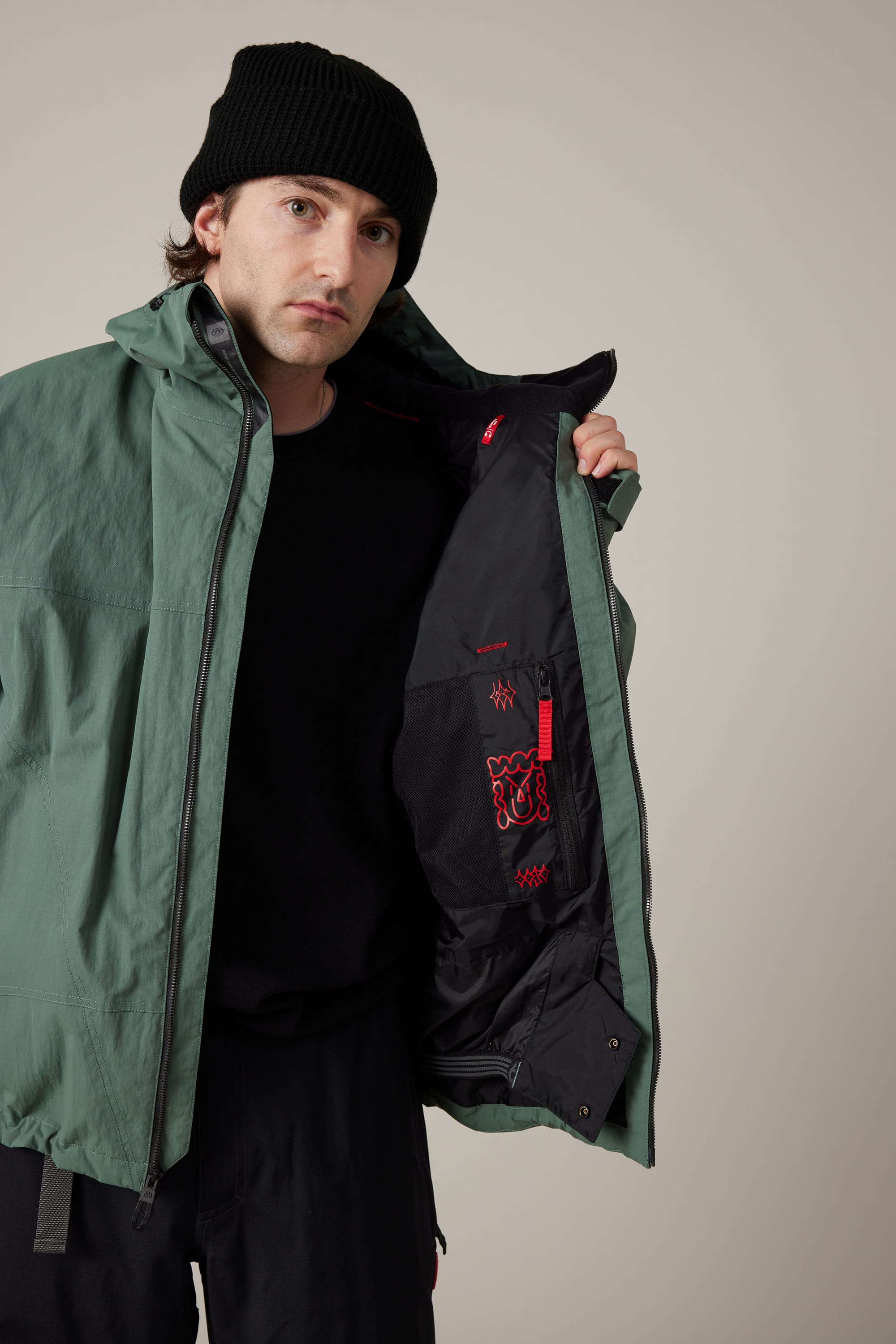 A person in a green 686 Men’s 686 Dojo® Shell Jacket and a black beanie opens their jacket to reveal the black and red interior lining, standing against a plain beige background.