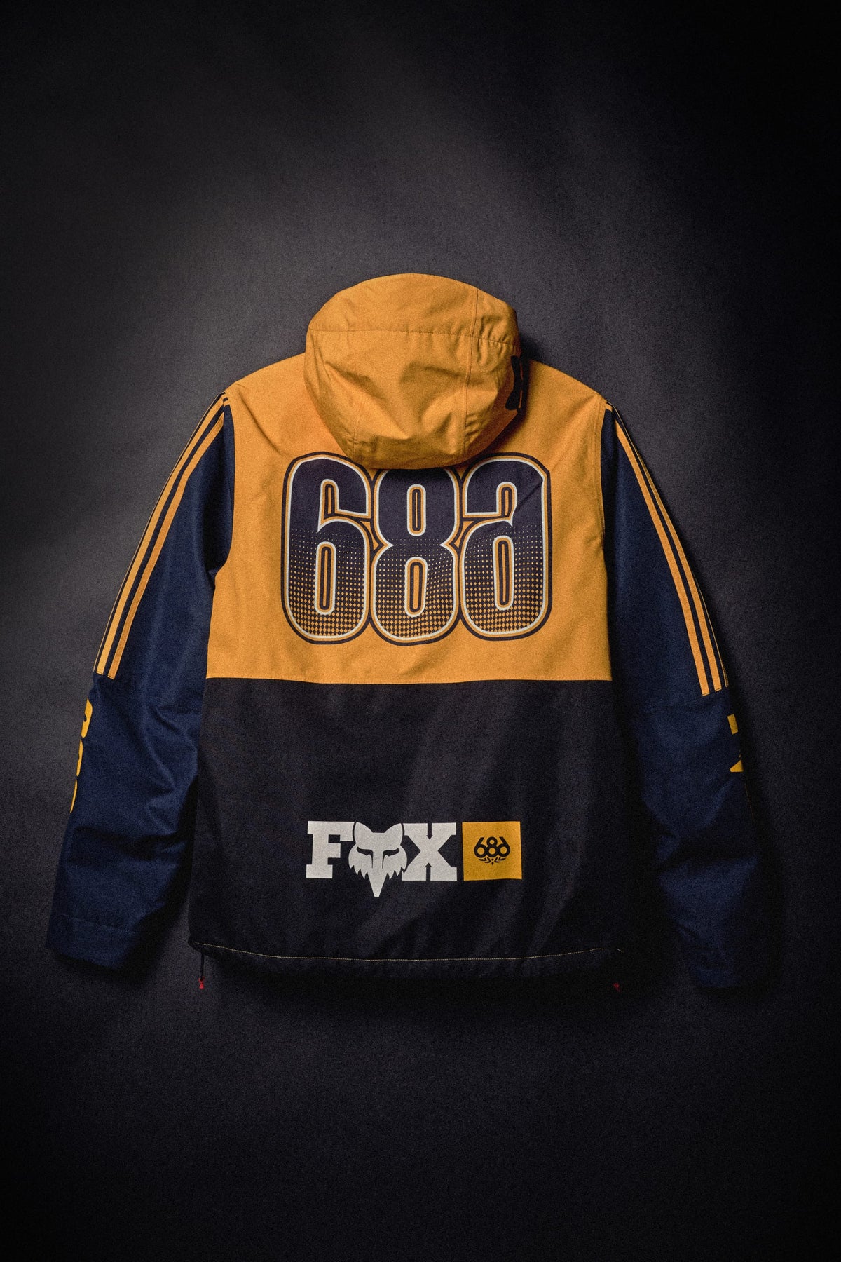 A gold and navy blue 686 Men’s Fox Racing Spec Insulated Jacket with 686 printed on the back and a Fox logo at the hem.