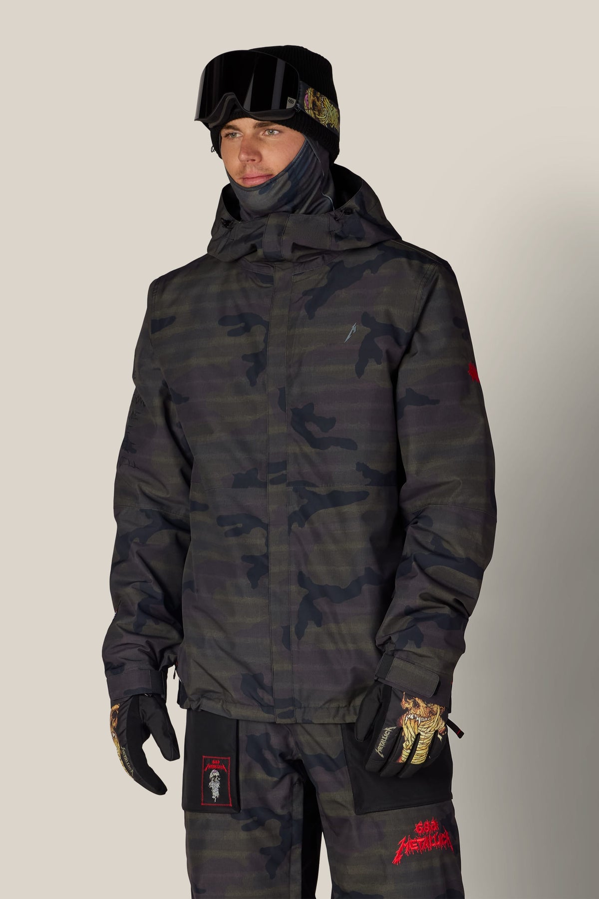 A person wearing a dark camouflage 686 Men’s Metallica One Insulated Jacket, matching pants, graphic design gloves, a neck gaiter, and goggles on their helmet.