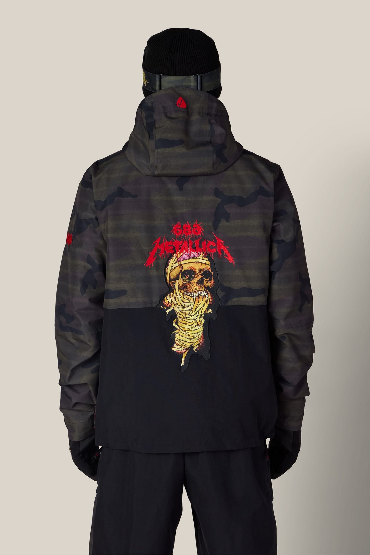 Back view of a person wearing a dark camouflage and black 686 Men’s Metallica One Insulated Jacket, featuring a Metallica mummy skull graphic on the back.