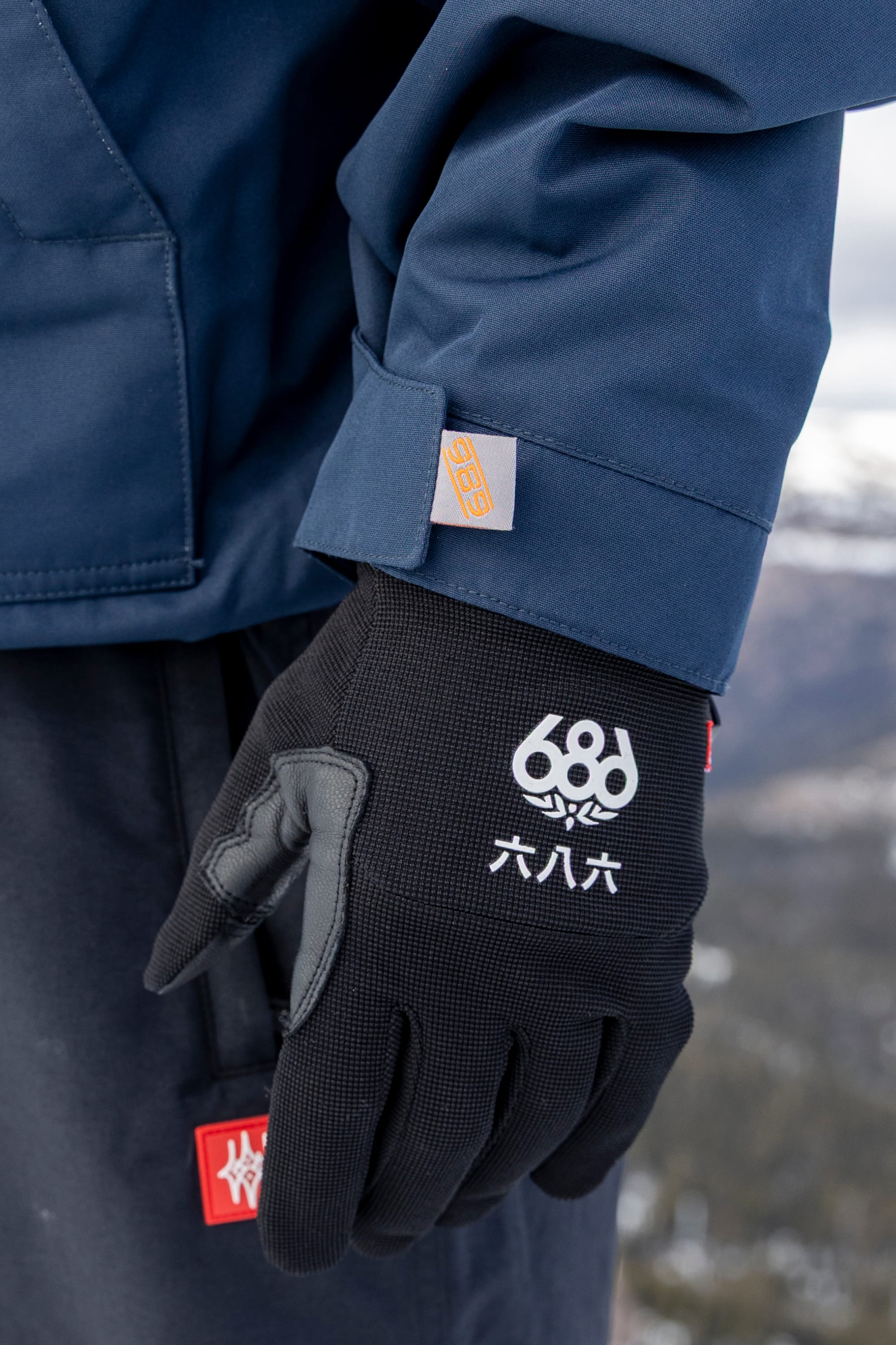 A person wearing the 686 Men’s 1994 Liberty Jacket in blue stands outdoors, paired with black 686 gloves featuring white Chinese characters, set against a blurred snowy landscape.