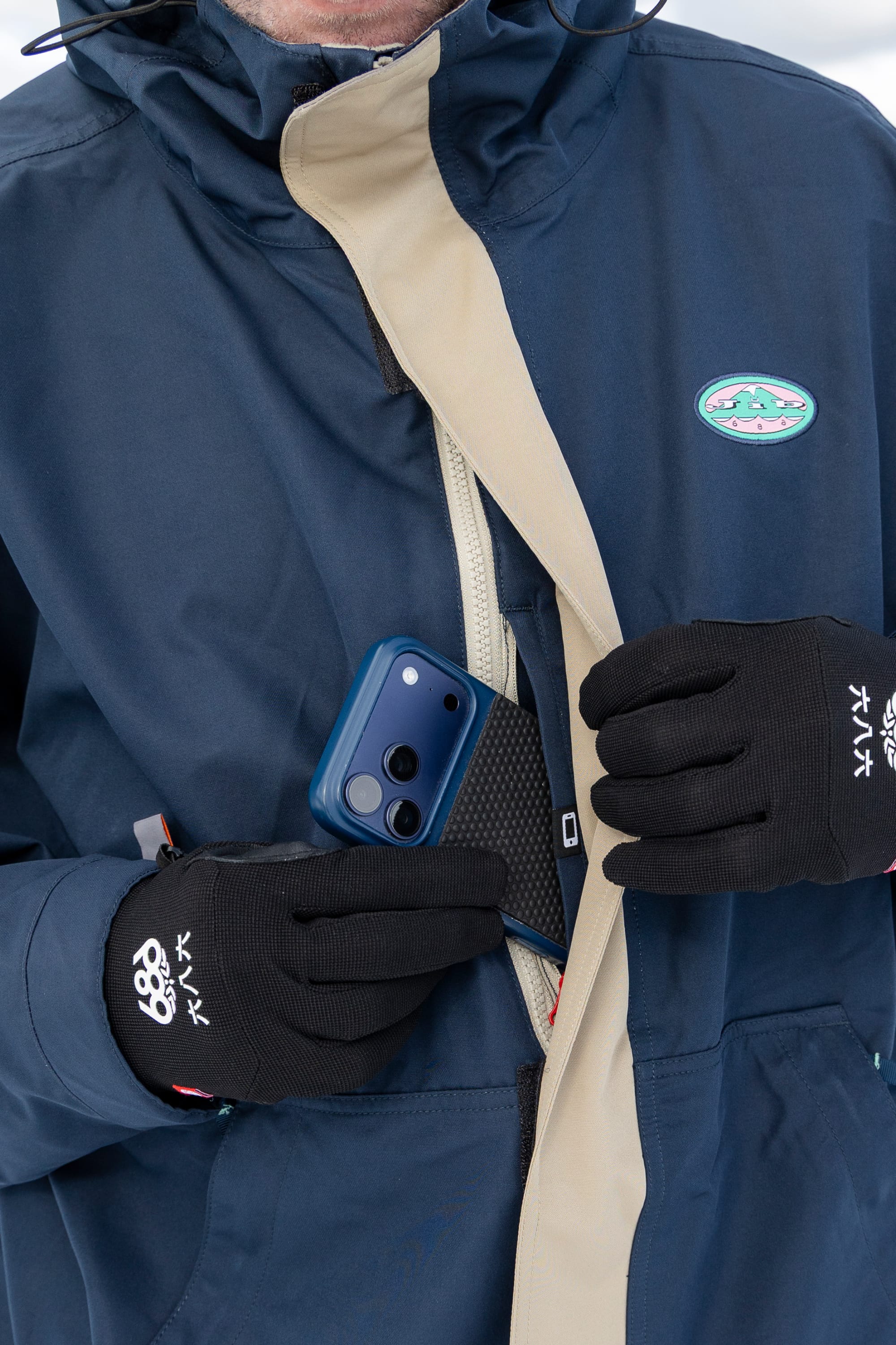 A person wearing black gloves and a blue 686 Men’s 1994 Liberty Jacket unzips a pocket to place or retrieve a blue smartphone. Only their hands and torso are visible; the face is not shown.