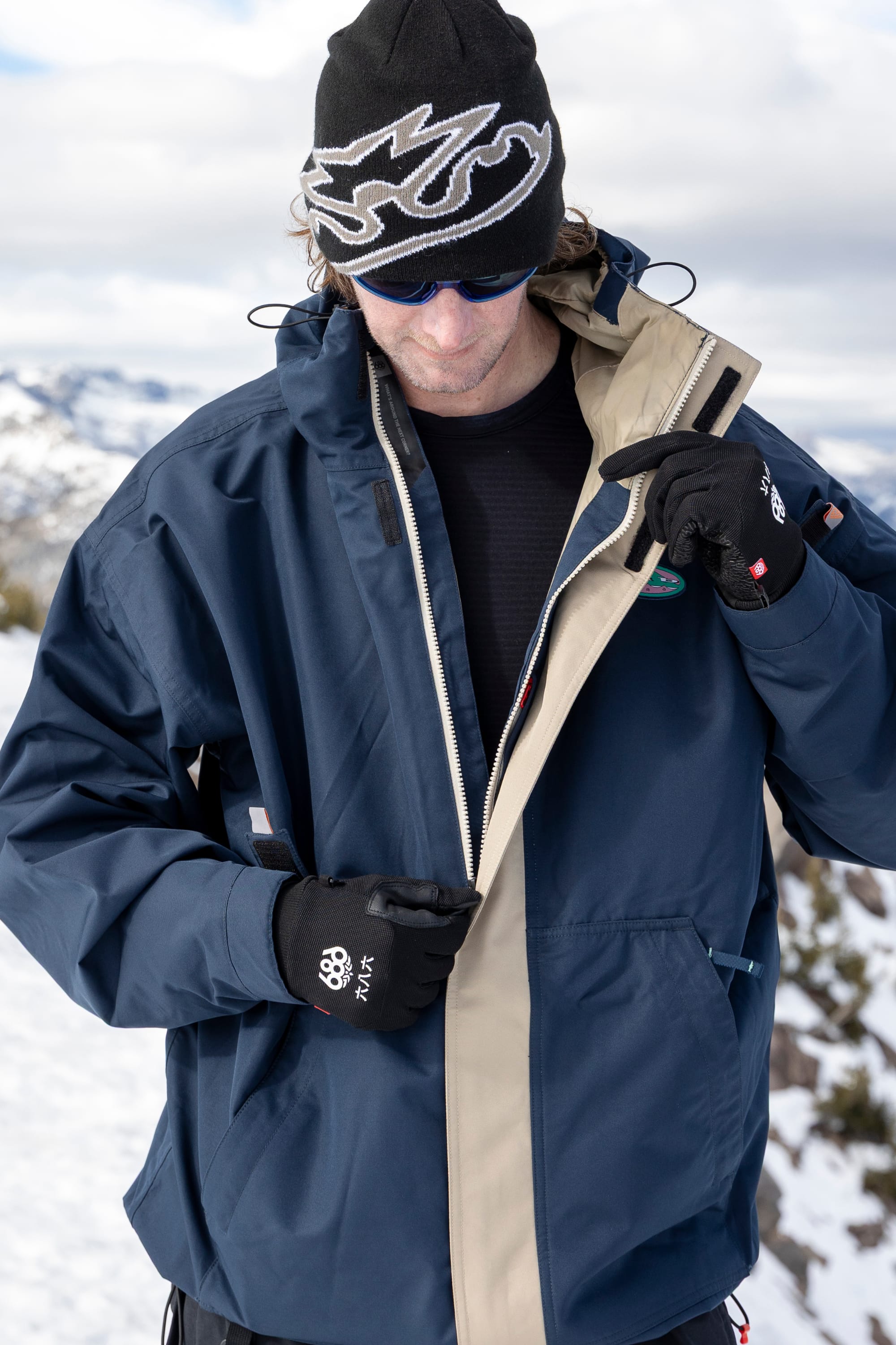 A person wearing the 686 Men’s 1994 Liberty Jacket, along with black gloves, sunglasses, and a beanie, stands outdoors in a snowy landscape, adjusting their jacket zipper.
