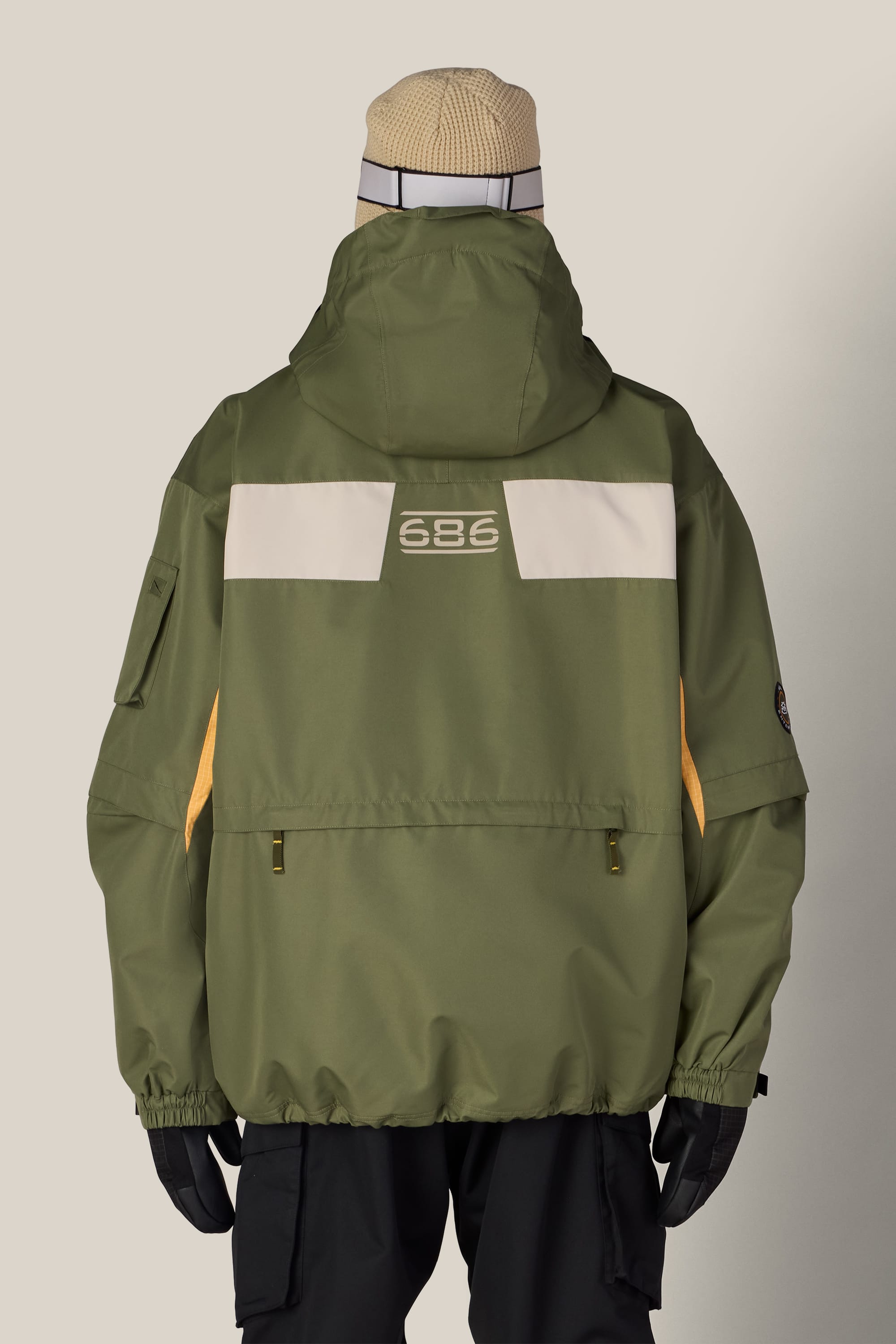A person wearing the 686 Men’s 2003 Atom Jacket in olive green, featuring a white stripe and the 686 logo on the back, along with black gloves, black pants, and a beige beanie, stands with their back to the camera.