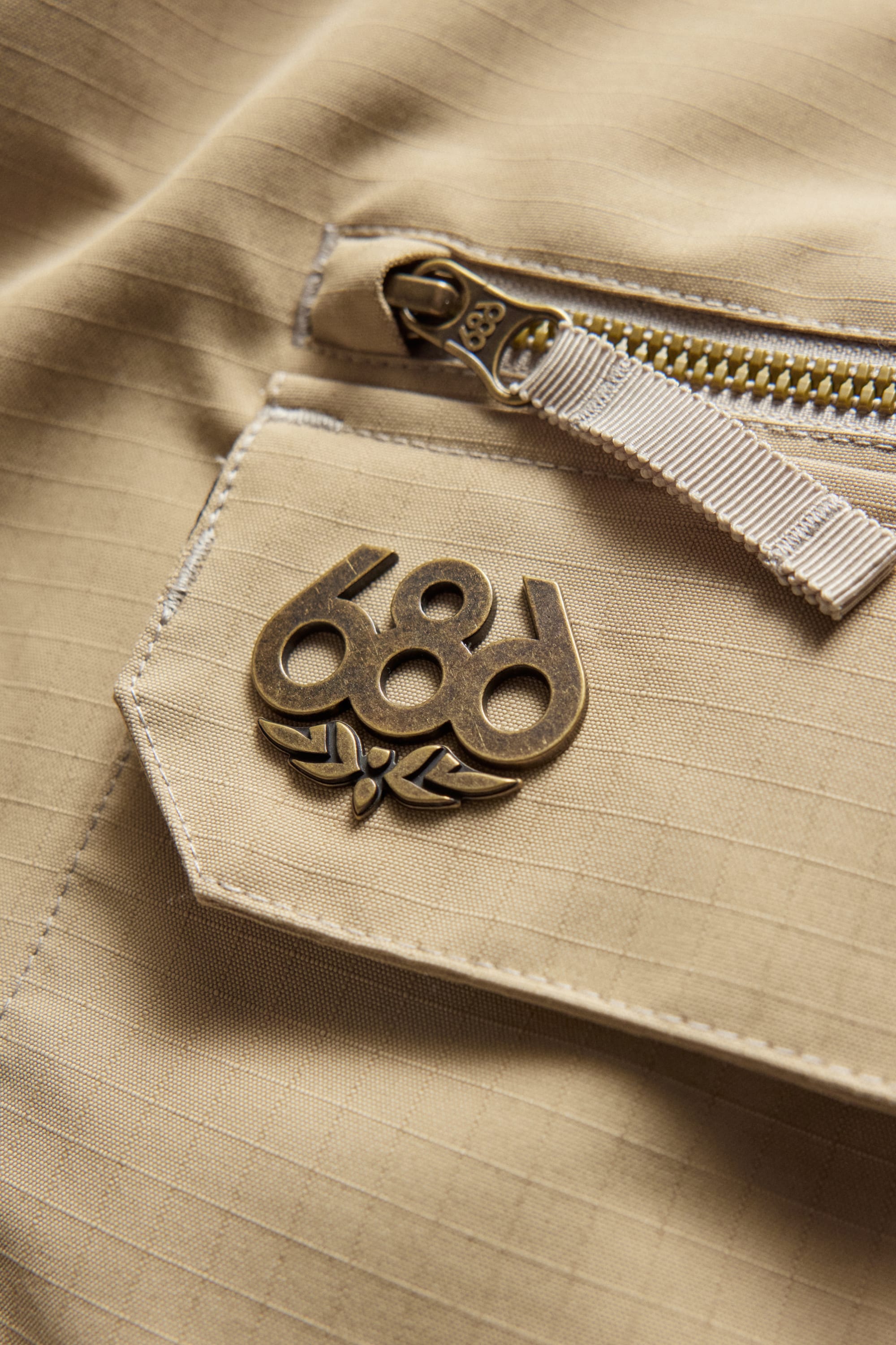 A close-up of the 686 Men’s Nightline Cargo Pant in beige showcases a brass 686 logo on the fabric pocket and a zippered flap, emphasizing the pant’s thoughtful design details. The zipper includes a matching beige pull tab, while the subtle grid pattern highlights the durable, waterproof polyester construction from 686.