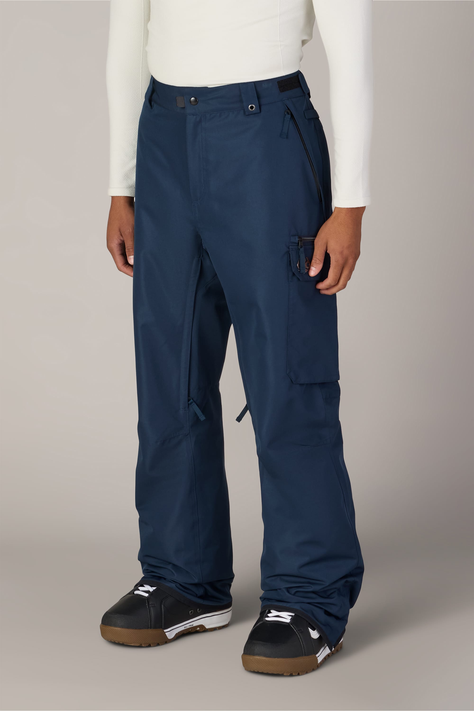 A person wearing 686 Men’s Nightline Cargo Pant in navy blue polyester, paired with a white long-sleeve shirt and black winter boots, stands against a plain light background.