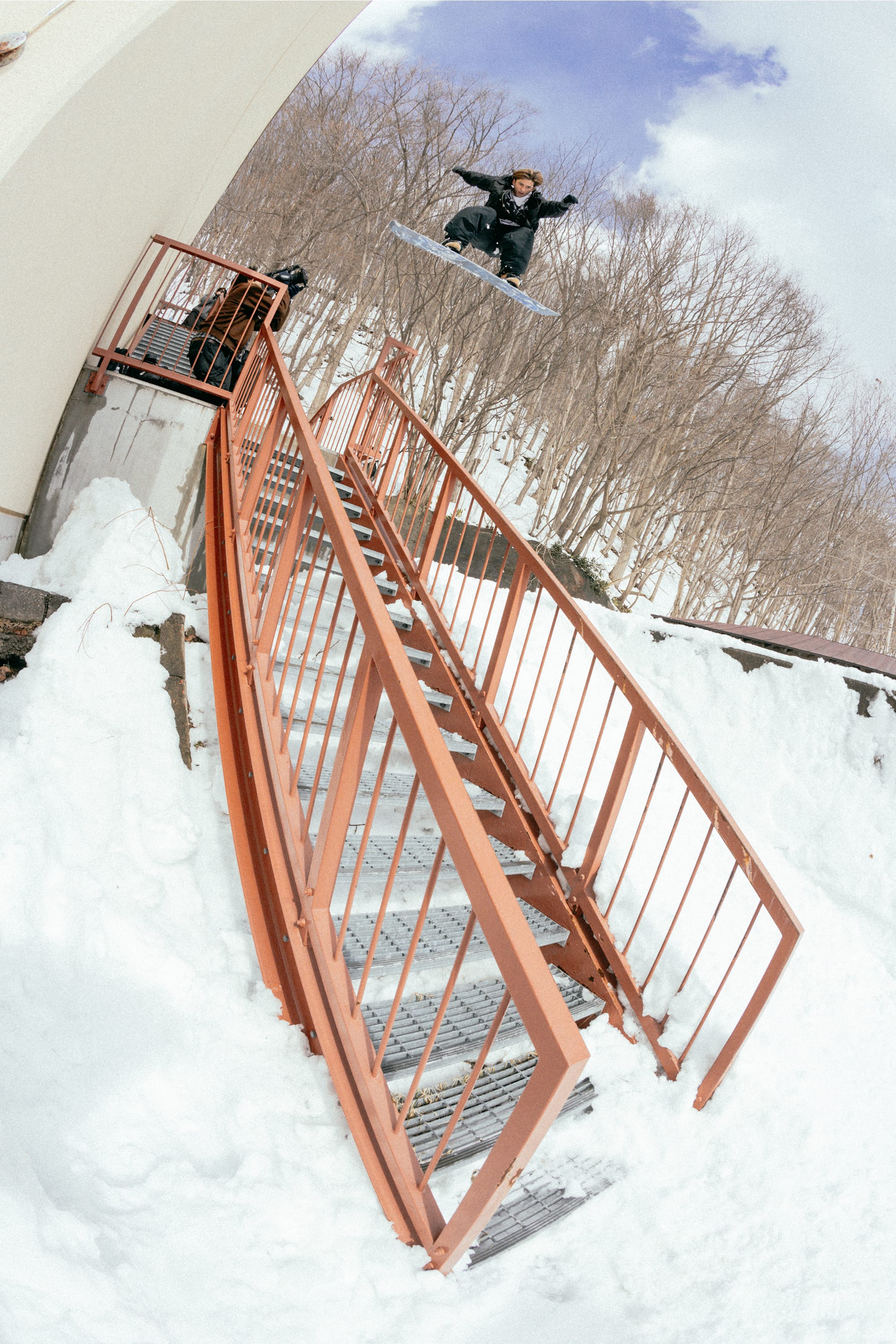 A snowboarder wearing the 686 Men’s 686 Dojo® GORE-TEX® Shell Pant by 686 performs a high jump off a snowy ledge over a red railing and stairs, surrounded by snow and bare trees under a partly cloudy sky.