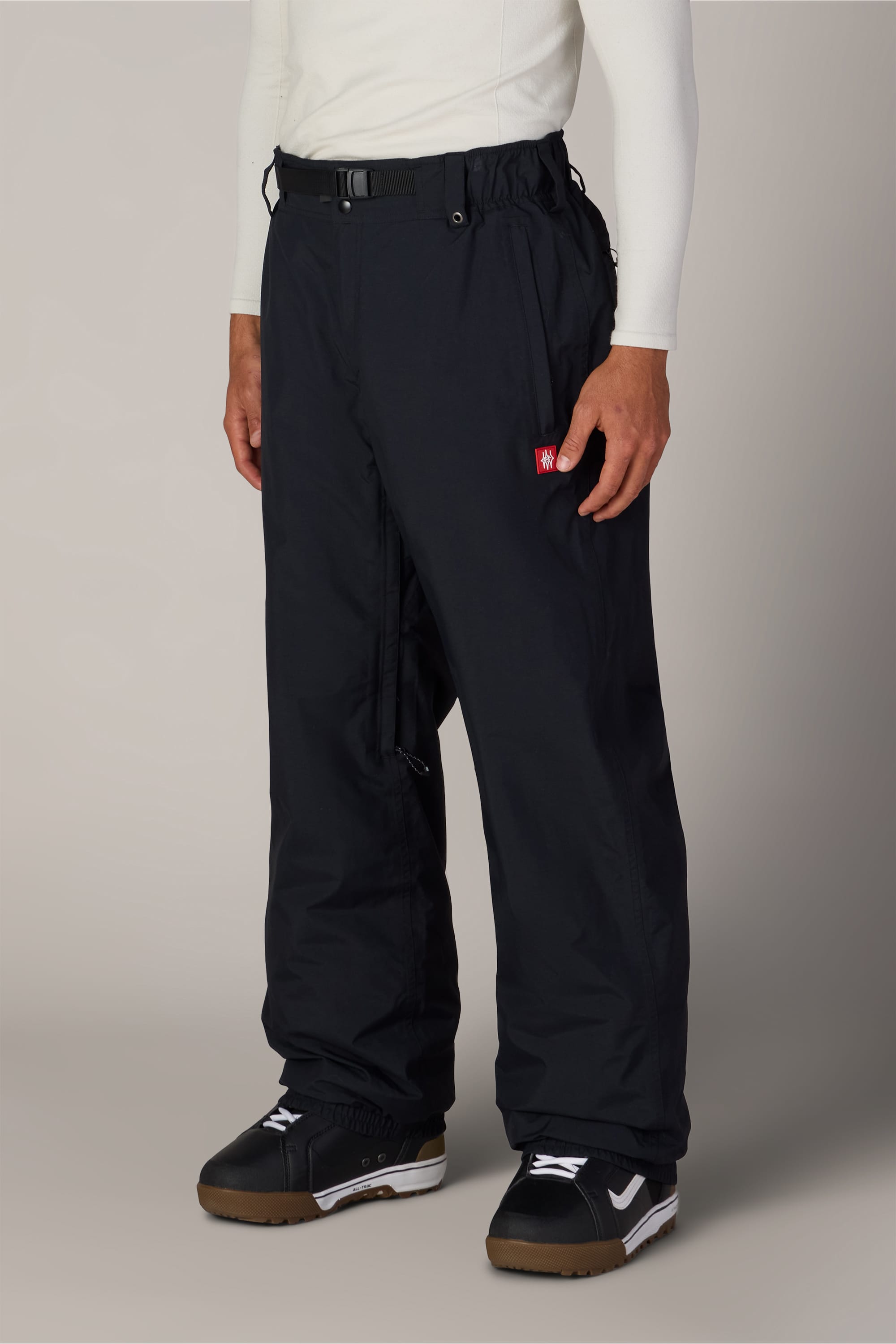 A person wearing the 686 Men’s Dojo® GORE-TEX® Shell Pant by 686, paired with black and white sneakers and a white long-sleeve shirt, stands against a plain background.