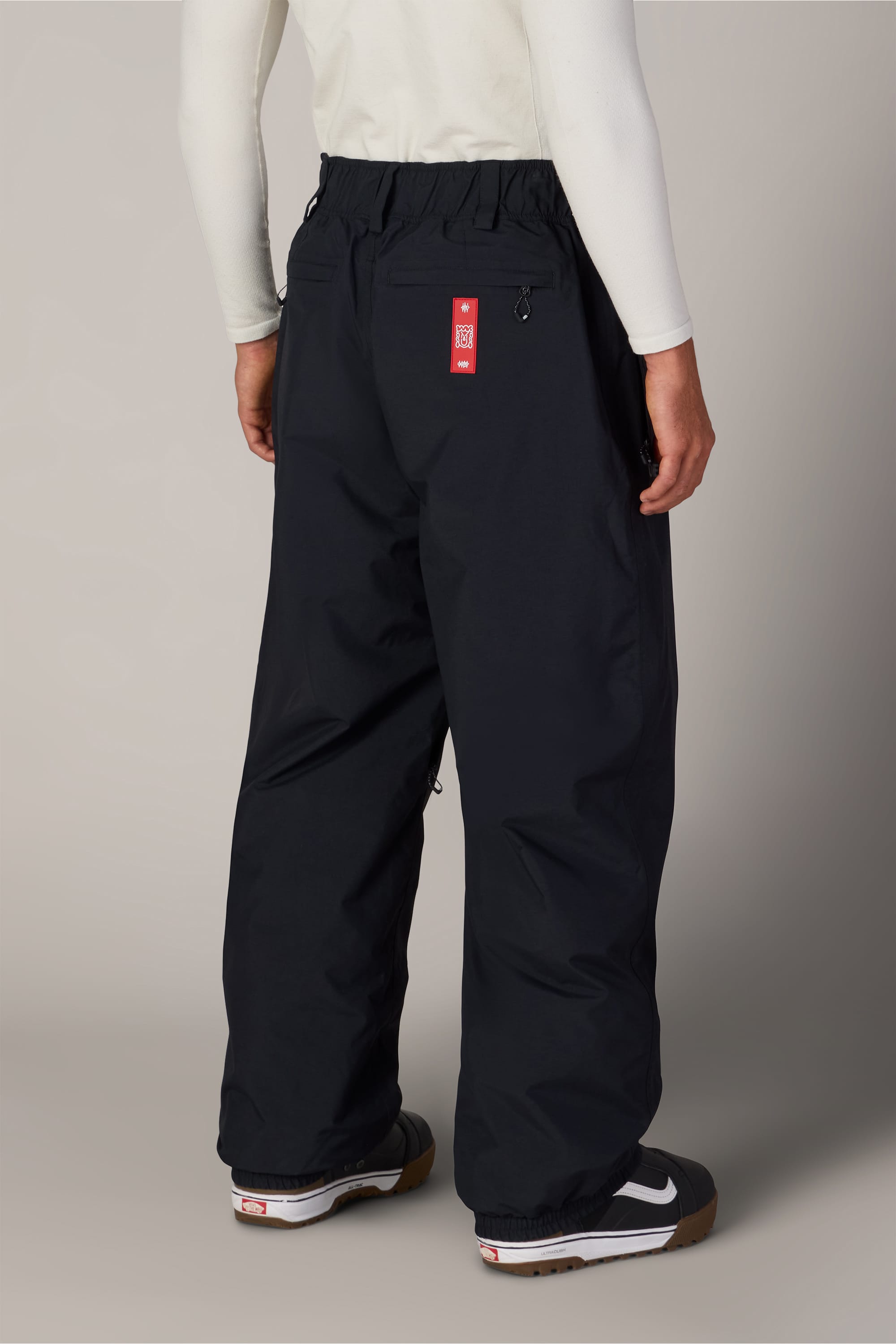 A person is seen from behind wearing the 686 Men’s 686 Dojo® GORE-TEX® Shell Pant by 686 in black, which offers a relaxed fit and features a red tag on the back waistband. They pair the pants with a white long-sleeve shirt and black sneakers with white soles.