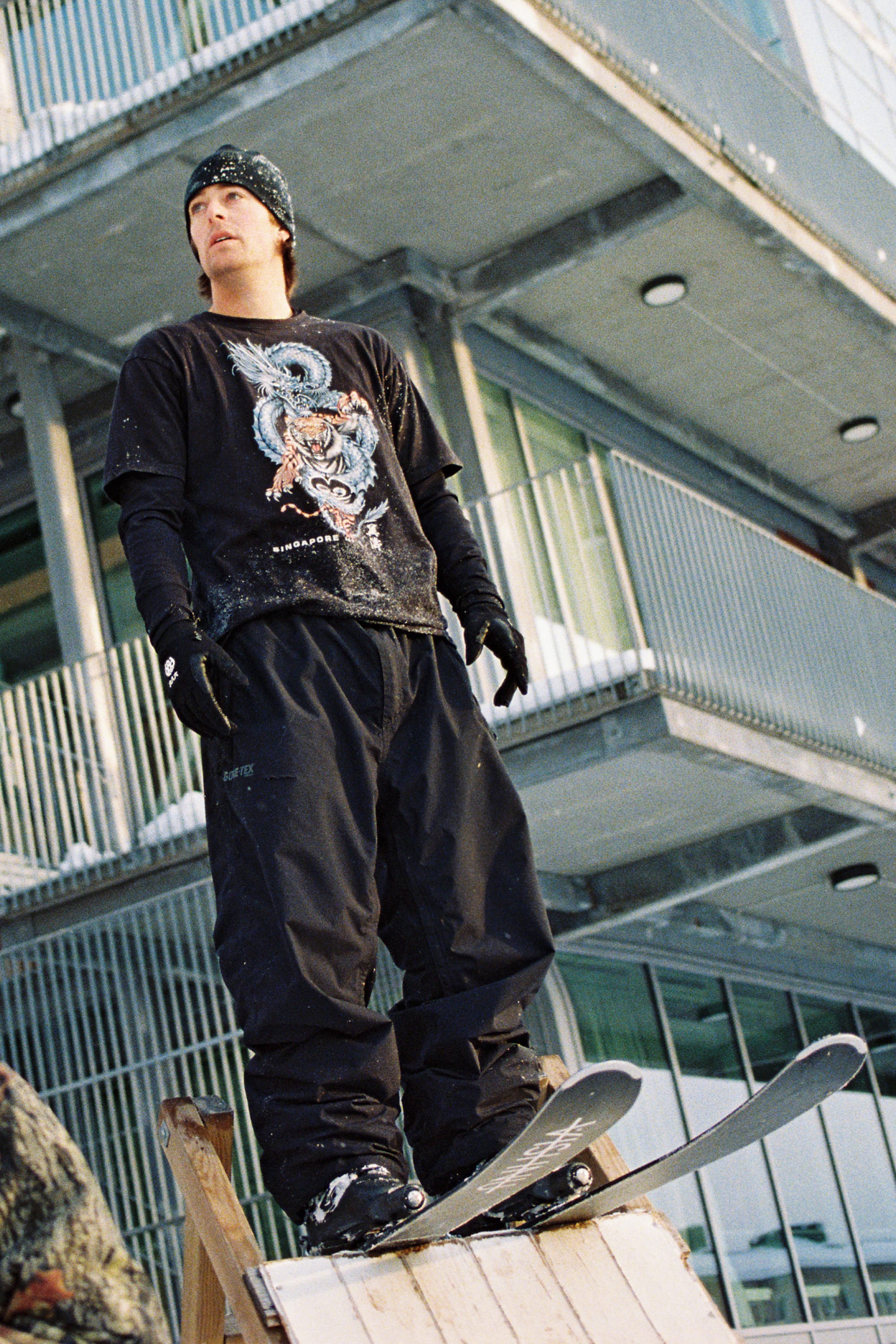 A snowboarder wearing a black beanie, gloves, and the 686 Men’s 686 Dojo® GORE-TEX® Shell Pant by 686 stands on a wooden ramp in front of a modern building, ready to ride.