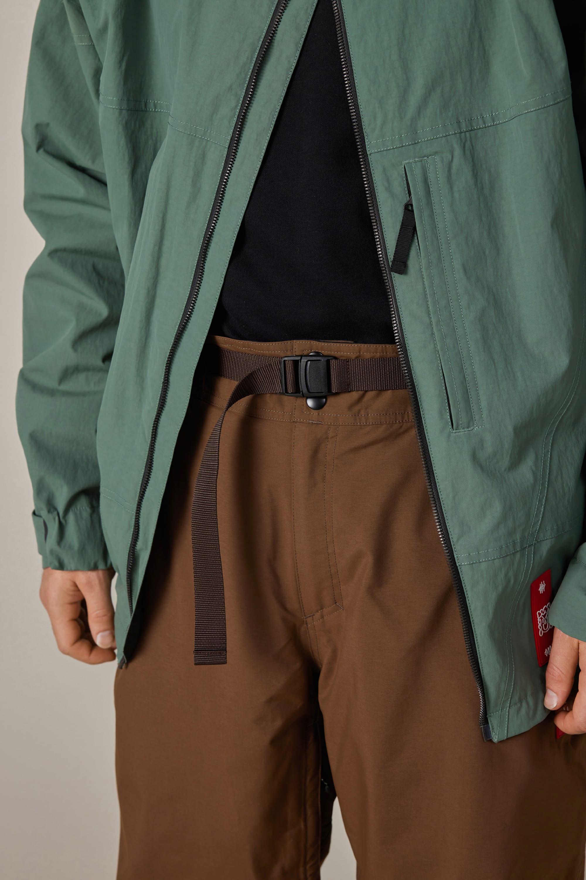 A person wearing a green jacket over a black shirt, paired with 686 Men’s 686 Dojo® GORE-TEX® Shell Pants in brown and accessorized with a black belt; only the midsection is visible.