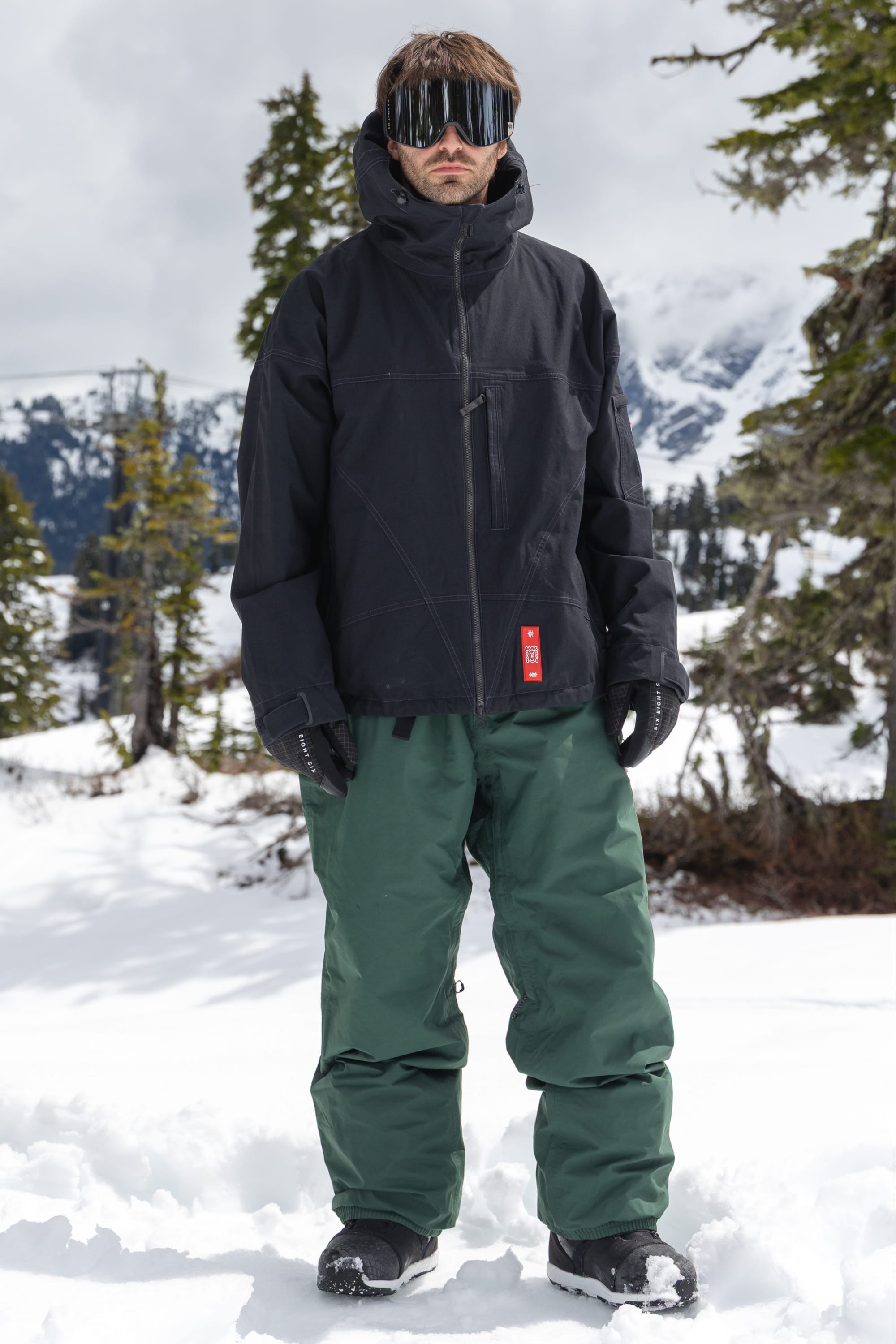 A person stands in the snow wearing a black jacket, black gloves, reflective ski goggles, and the 686 Men’s 686 Dojo® GORE-TEX® Shell Pant by 686, with snow-covered trees and mountains in the background.