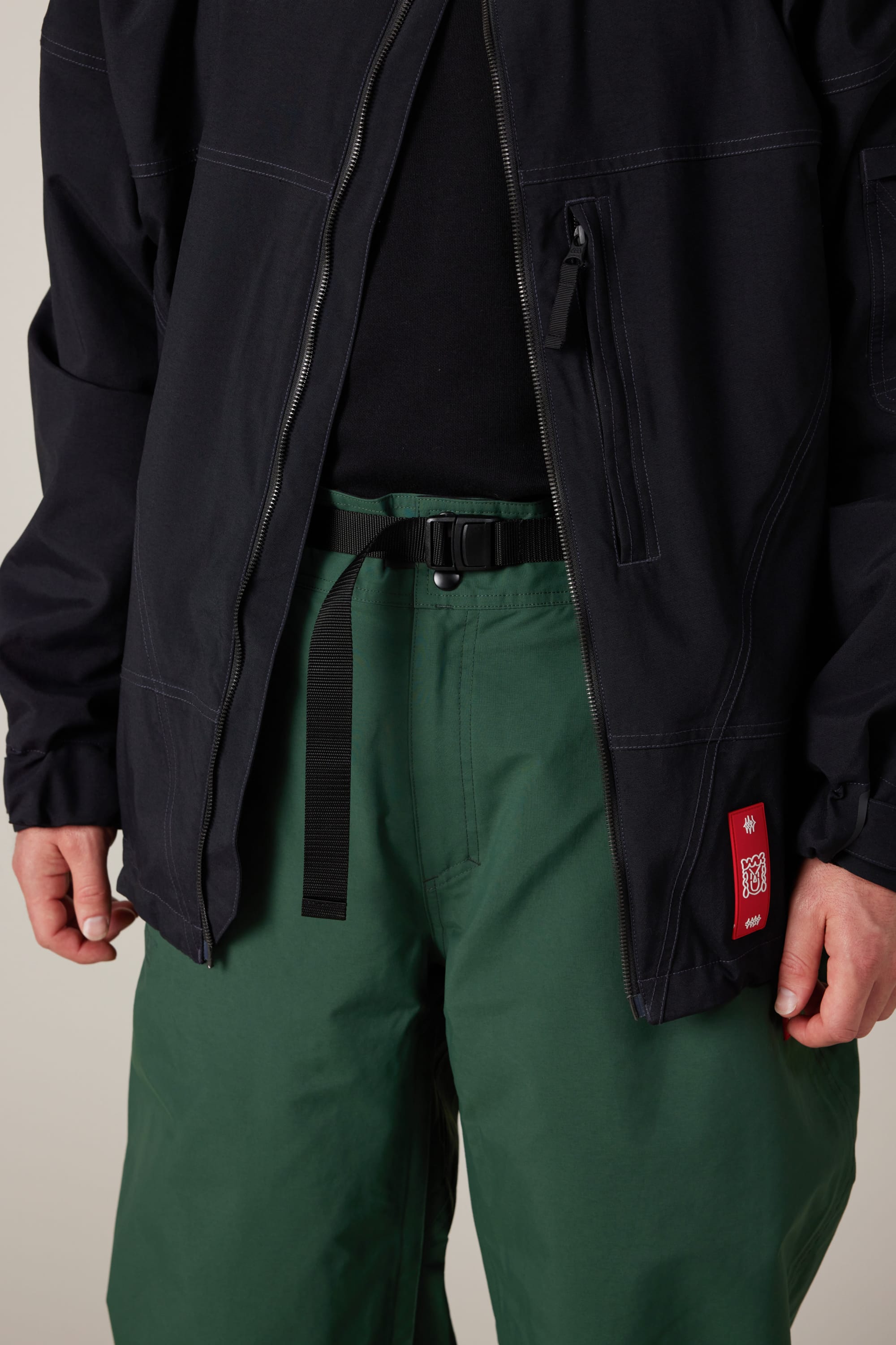 A person is shown from the torso to upper legs, wearing a black jacket with a visible zipper pocket and a small red patch near the hem, paired with a black shirt and green 686 Men’s Dojo® GORE-TEX® Shell Pants by 686, secured with a black belt.