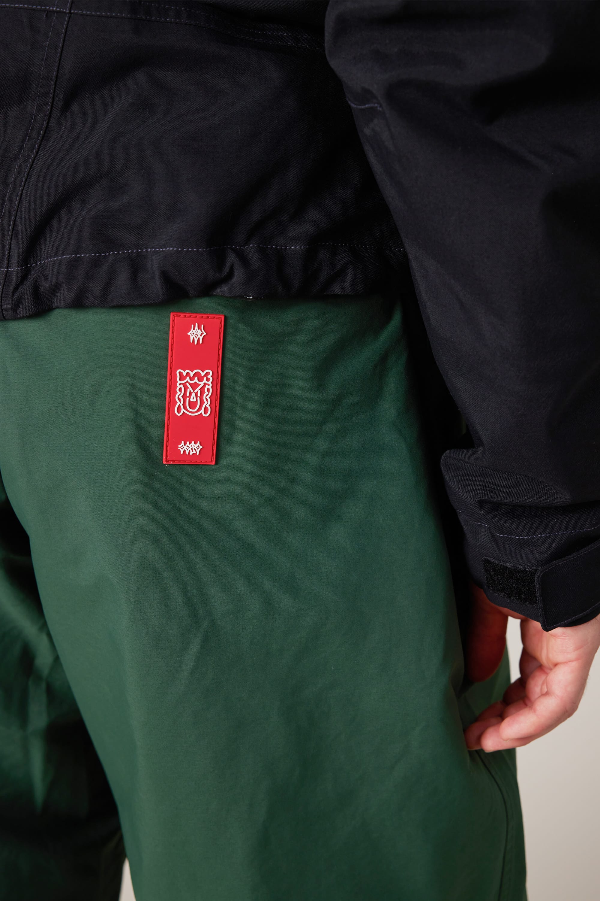 A person wearing green 686 Men’s 686 Dojo® GORE-TEX® Shell Pants and a dark jacket; a red tag with white designs is attached to the back of the pants near the waistband.