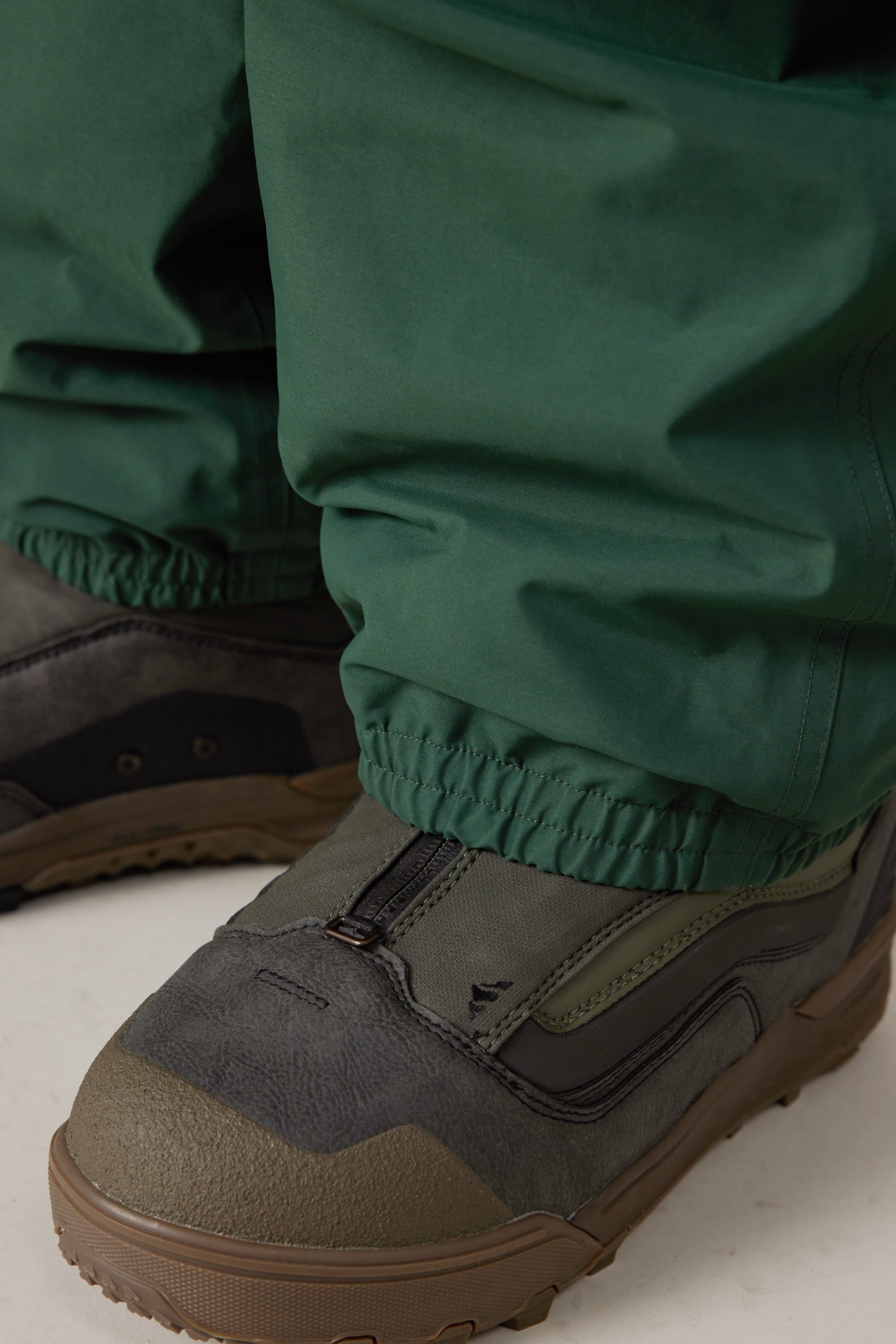 Close-up of a person wearing 686 Men’s 686 Dojo® GORE-TEX® Shell Pants by 686 with elastic cuffs over rugged, dark green and brown outdoor shoes on a light surface.
