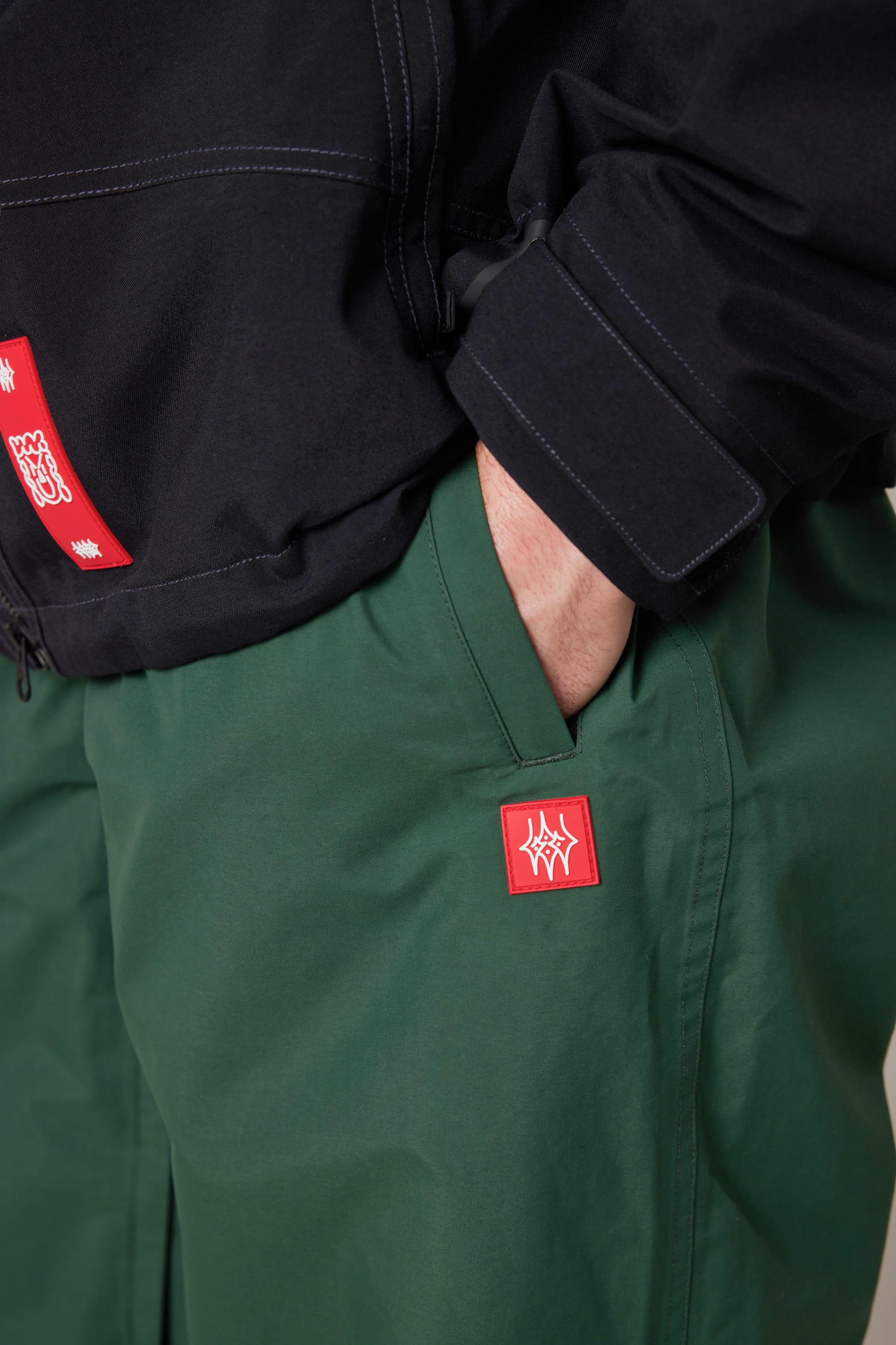 A person wearing the 686 Men’s Dojo® GORE-TEX® Shell Pant in green with their hand tucked into the pocket, paired with a black jacket featuring red and white patches.