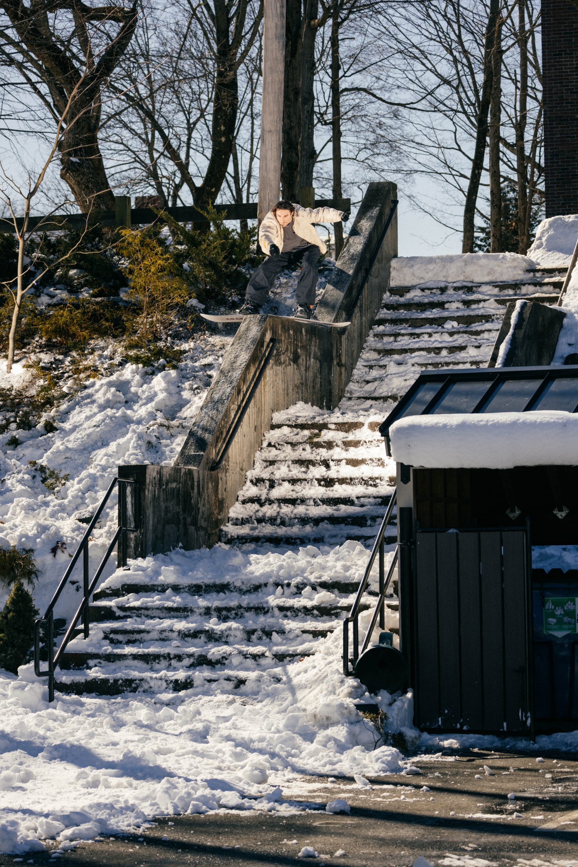Wearing the 686 Men’s 686 Dojo® Shell Pant, a snowboarder dressed in a light jacket rides down a snow-covered handrail beside outdoor stairs, with snow and bare trees around on a sunny winter day.