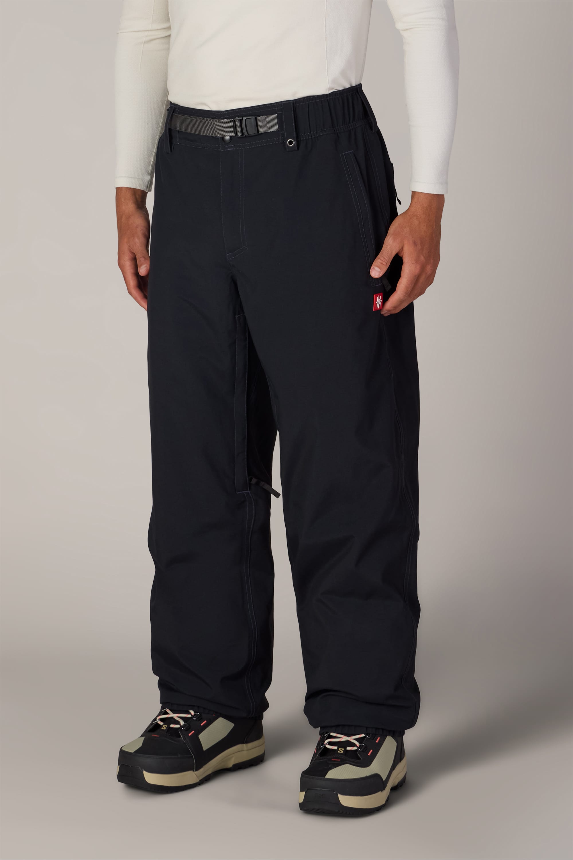 A person wearing a white long-sleeve shirt, black 686 Men’s 686 Dojo® Shell Pants with a belt, and black and beige hiking shoes stands facing forward against a plain background.