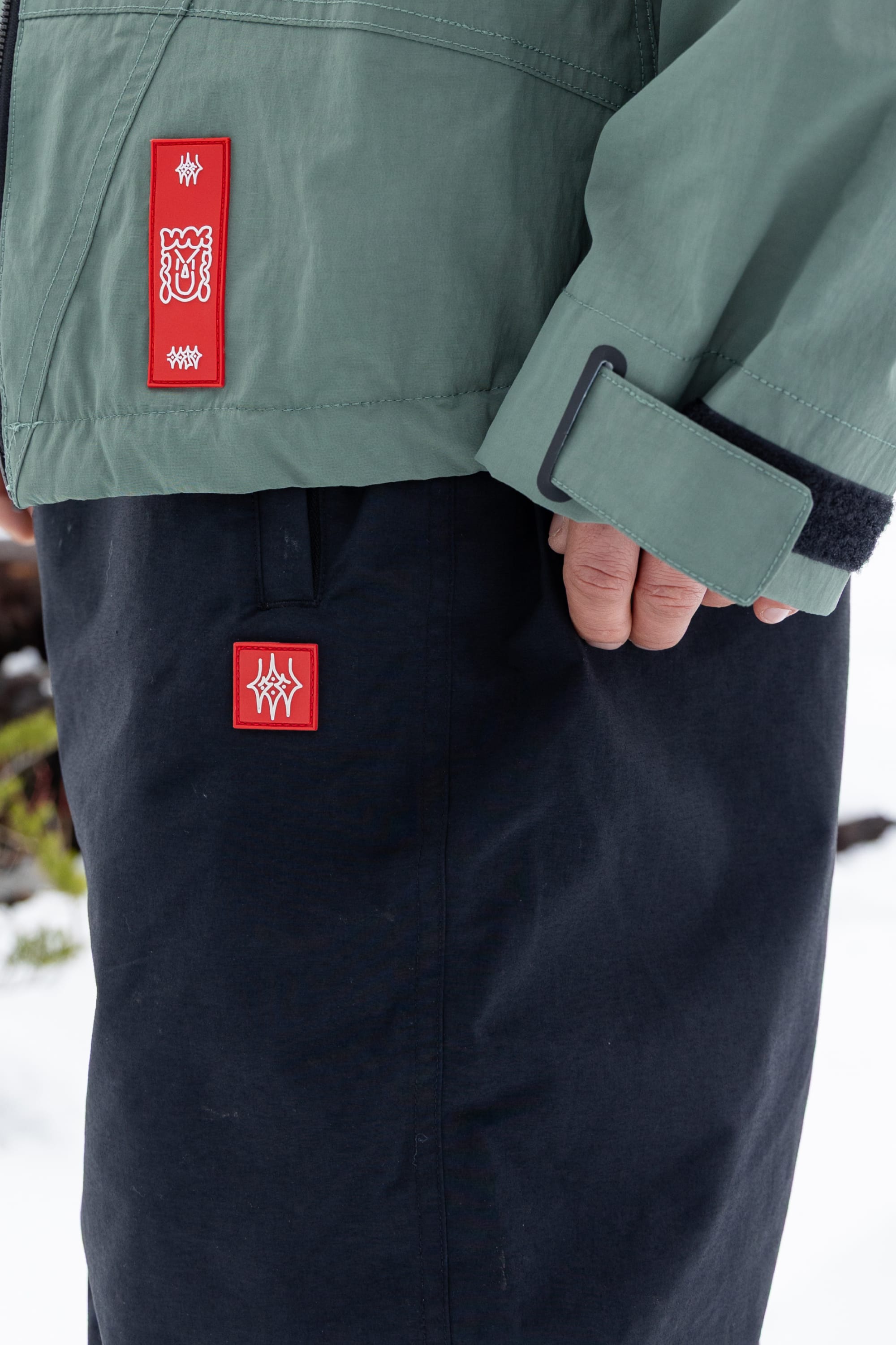 A person wearing a green jacket and the 686 Men’s 686 Dojo® Shell Pant, black nylon pants by 686 featuring red and white rectangular patches, stands outdoors on snow.