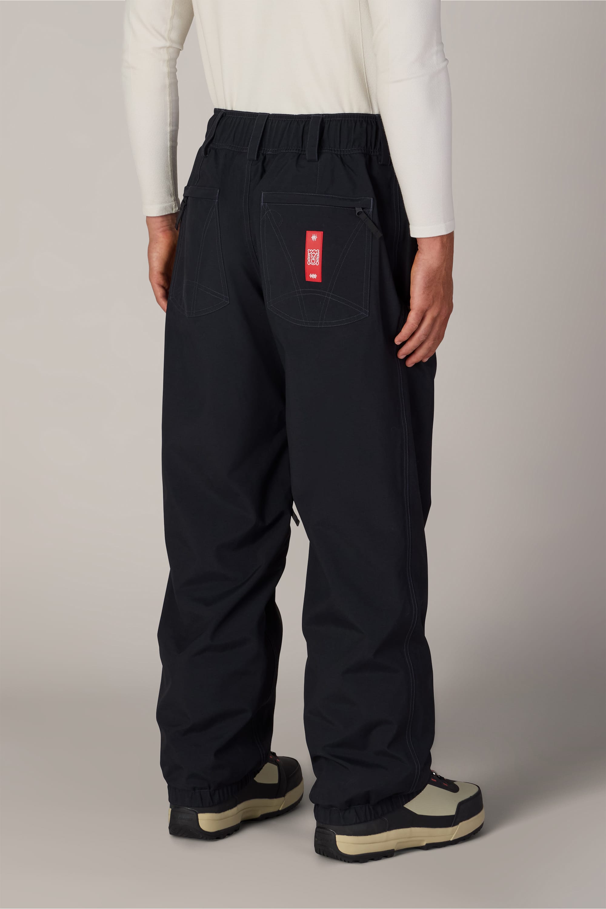 A person wearing loose black 686 Men’s 686 Dojo® Shell Pants by 686, featuring a red tag on the back pocket, paired with a white long-sleeve shirt and black and beige boots, is standing against a plain background, shown from the back.