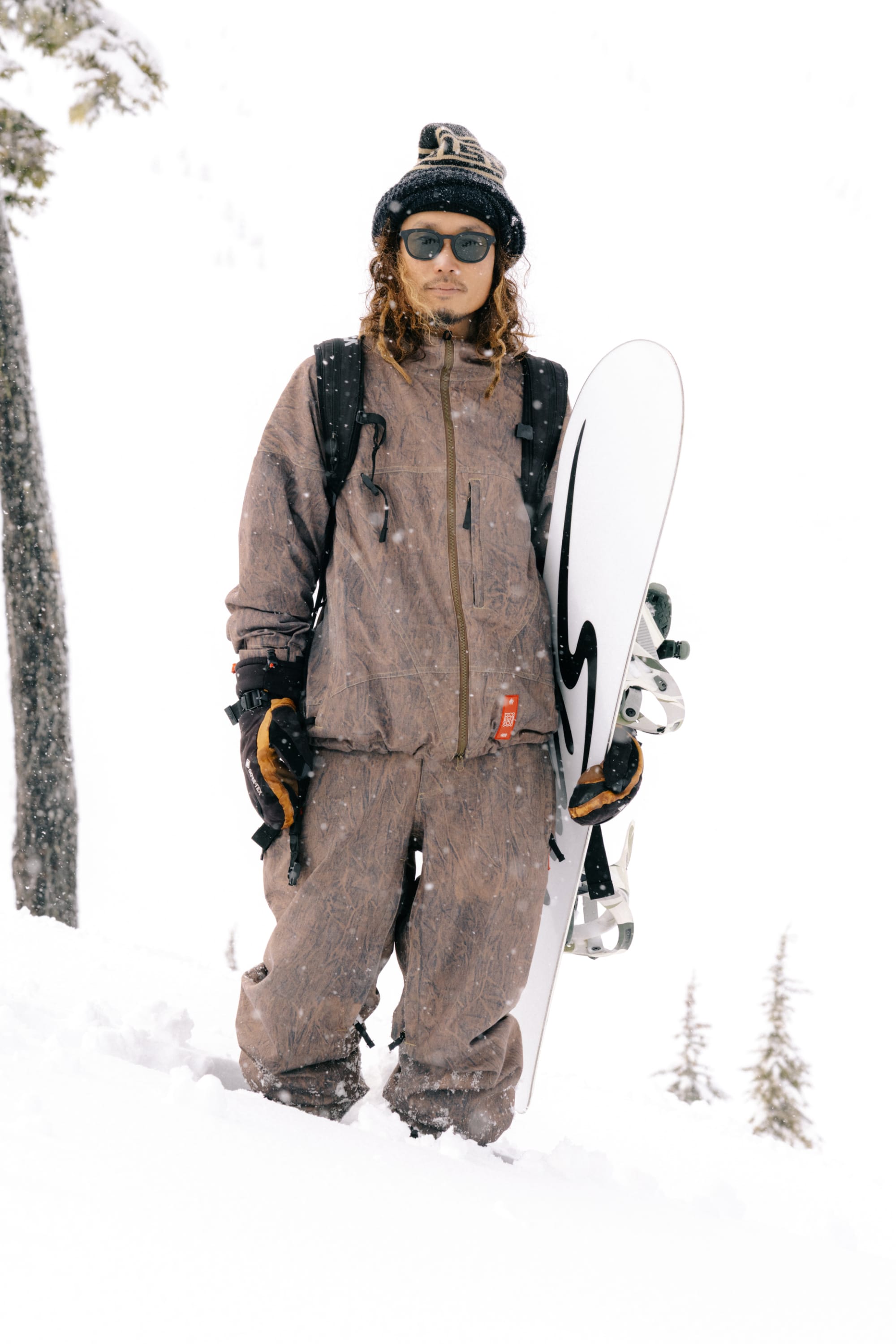 A person wearing brown snow gear, black gloves, and sunglasses stands in the snow holding a snowboard, with trees visible in the background. Their outfit features the 686 Men’s 686 Dojo® Shell Pant by 686, providing both style and durability for outdoor adventures.