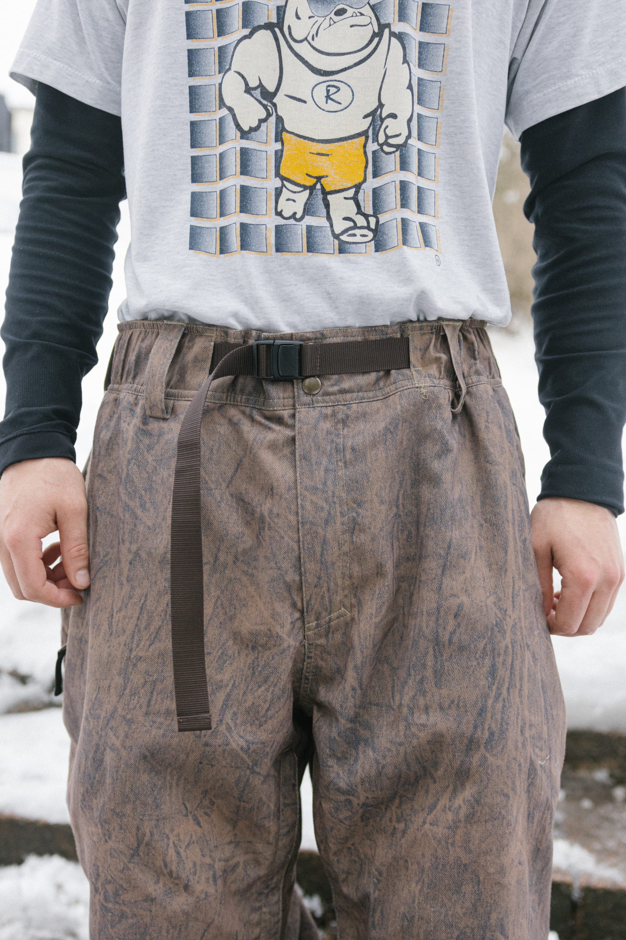 A person standing outdoors near snow is wearing 686 Men’s 686 Dojo® Shell Pants in a patterned brown color with a black belt, paired with a gray T-shirt featuring a cartoon dog in yellow shorts and a long-sleeved black shirt underneath.