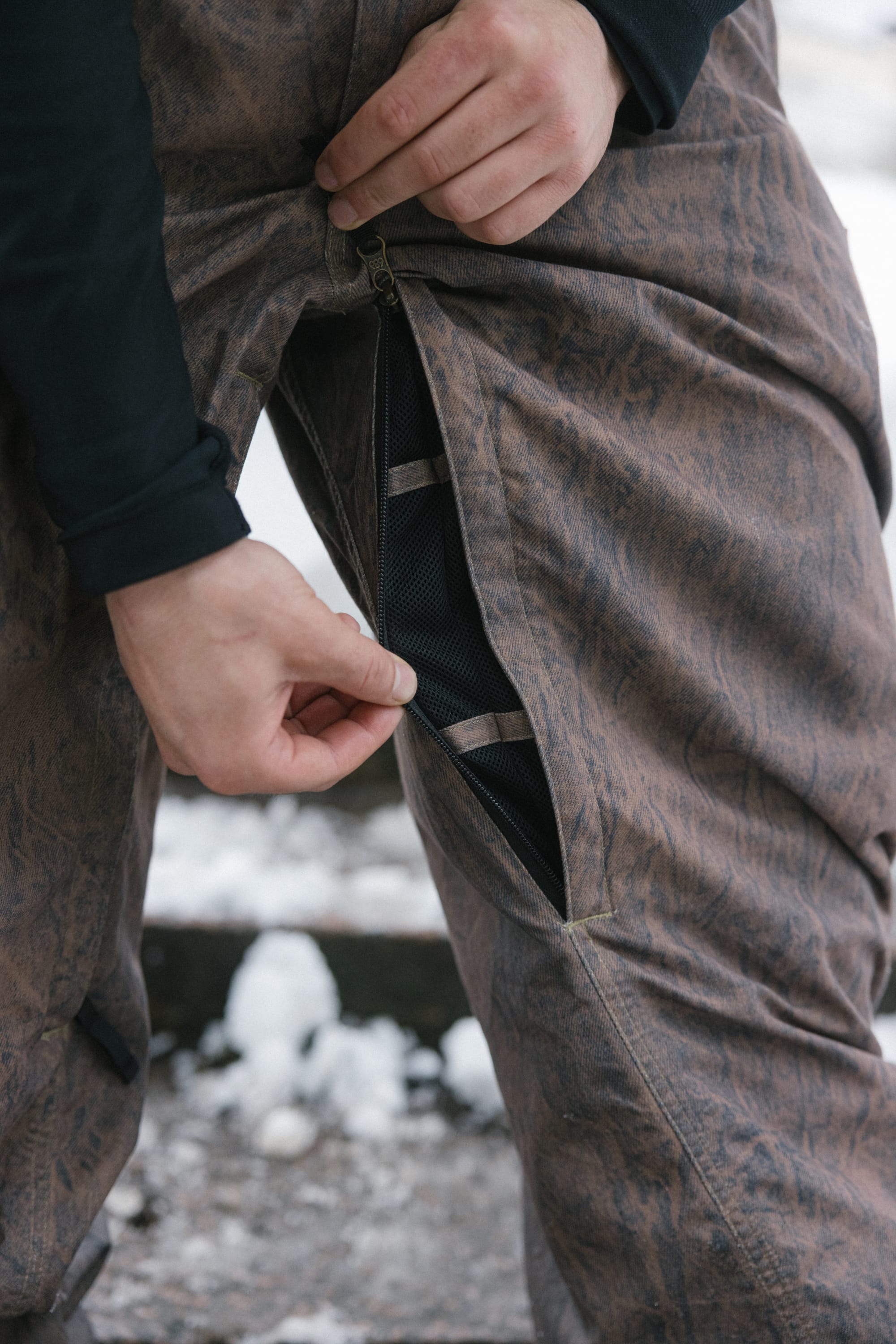 A person’s hand opens a zippered vent on the 686 Men’s Dojo® Shell Pant in brown, revealing a black mesh lining underneath. Snow and wooden steps are visible in the blurred background.