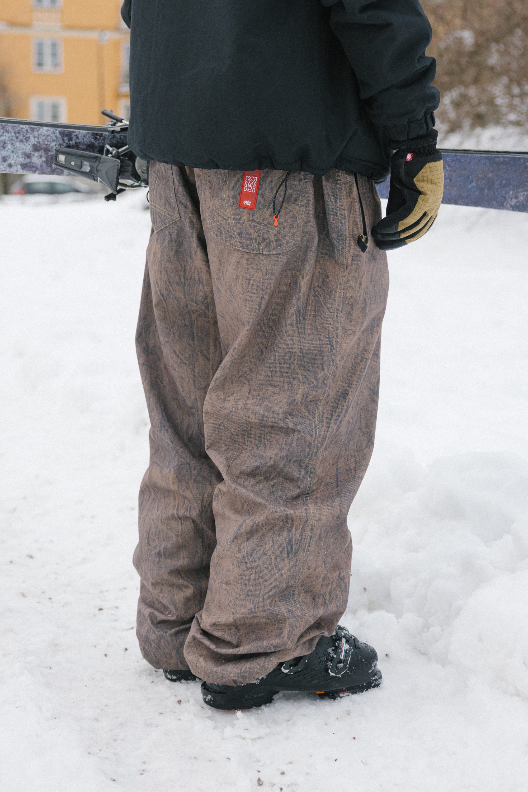 A person wearing baggy brown 686 Men’s Dojo® Shell Pants, along with a black jacket and gloves, is holding skis while standing on snow. The image is cropped above the waist, with a yellow building and trees visible in the background.