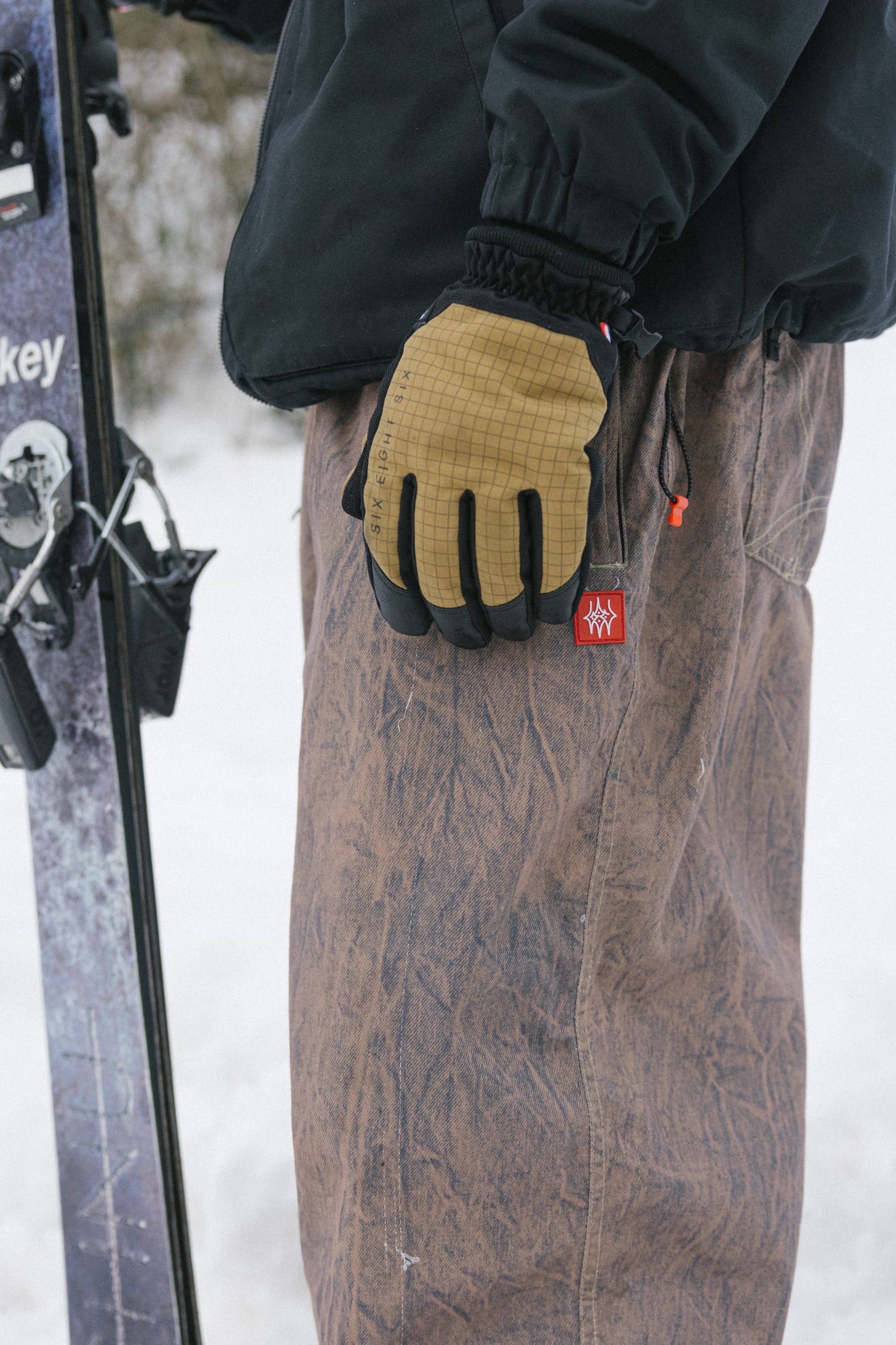 A person in the 686 Men’s 686 Dojo® Shell Pant stands in the snow holding skis, wearing tan gloves and a black jacket, with only their lower half visible.