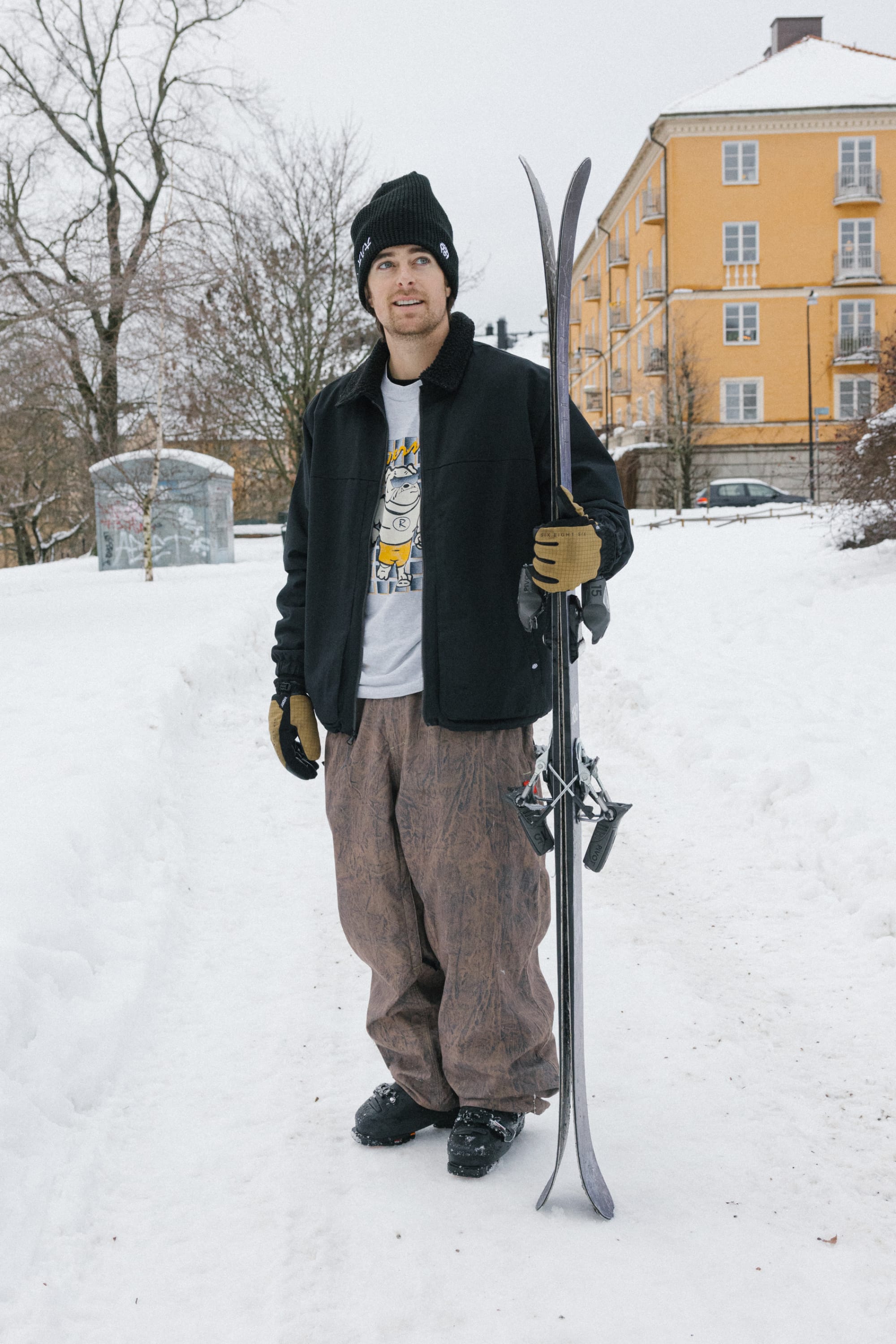 A person stands on a snowy path holding skis, wearing a black beanie, jacket, gloves, and the 686 Men’s 686 Dojo® Shell Pant by 686. An apartment building and trees are visible in the background.