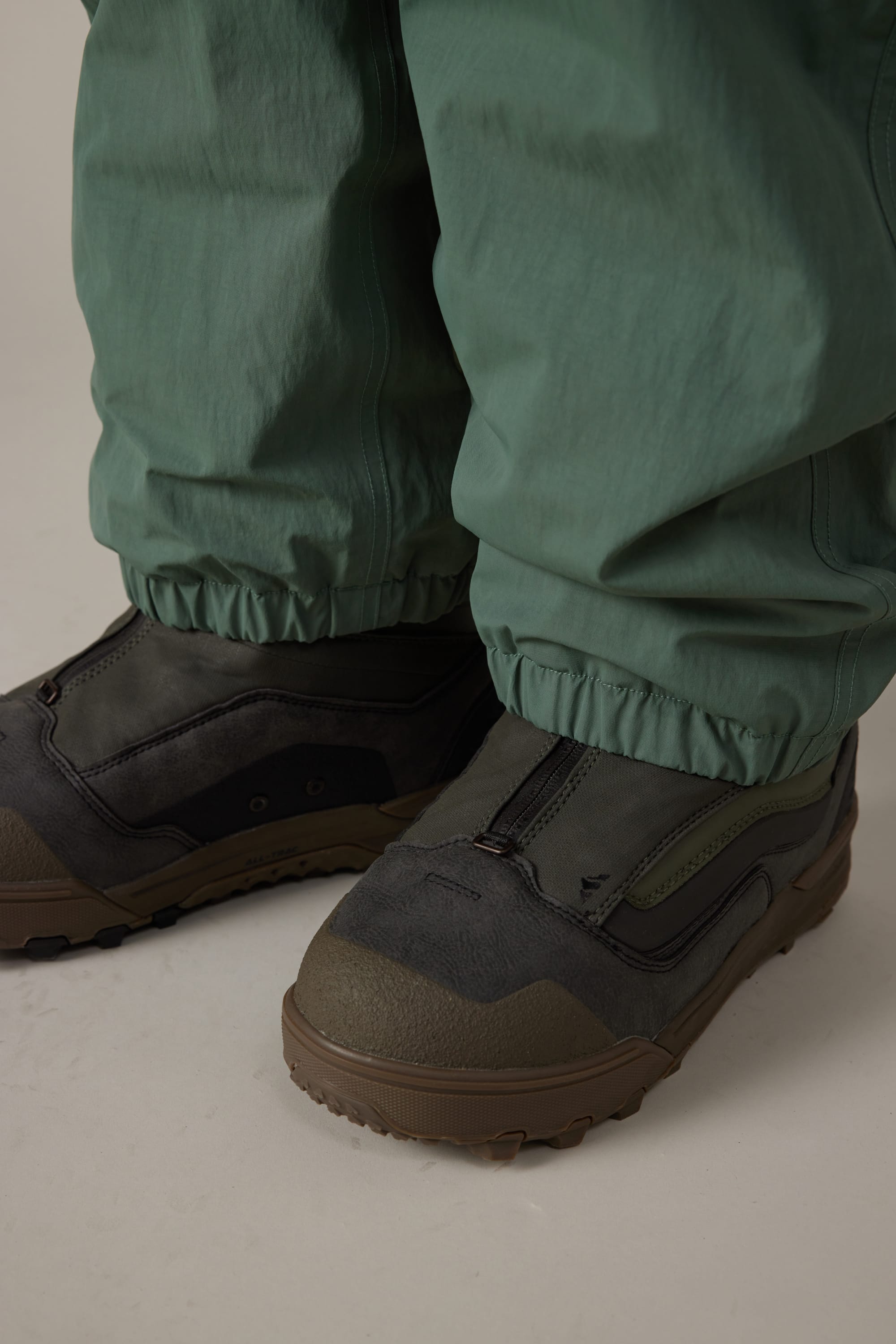 Close-up of a person wearing 686 Men’s 686 Dojo® Shell Pant in green with elastic cuffs and dark green outdoor boots with brown soles, standing on a light surface.