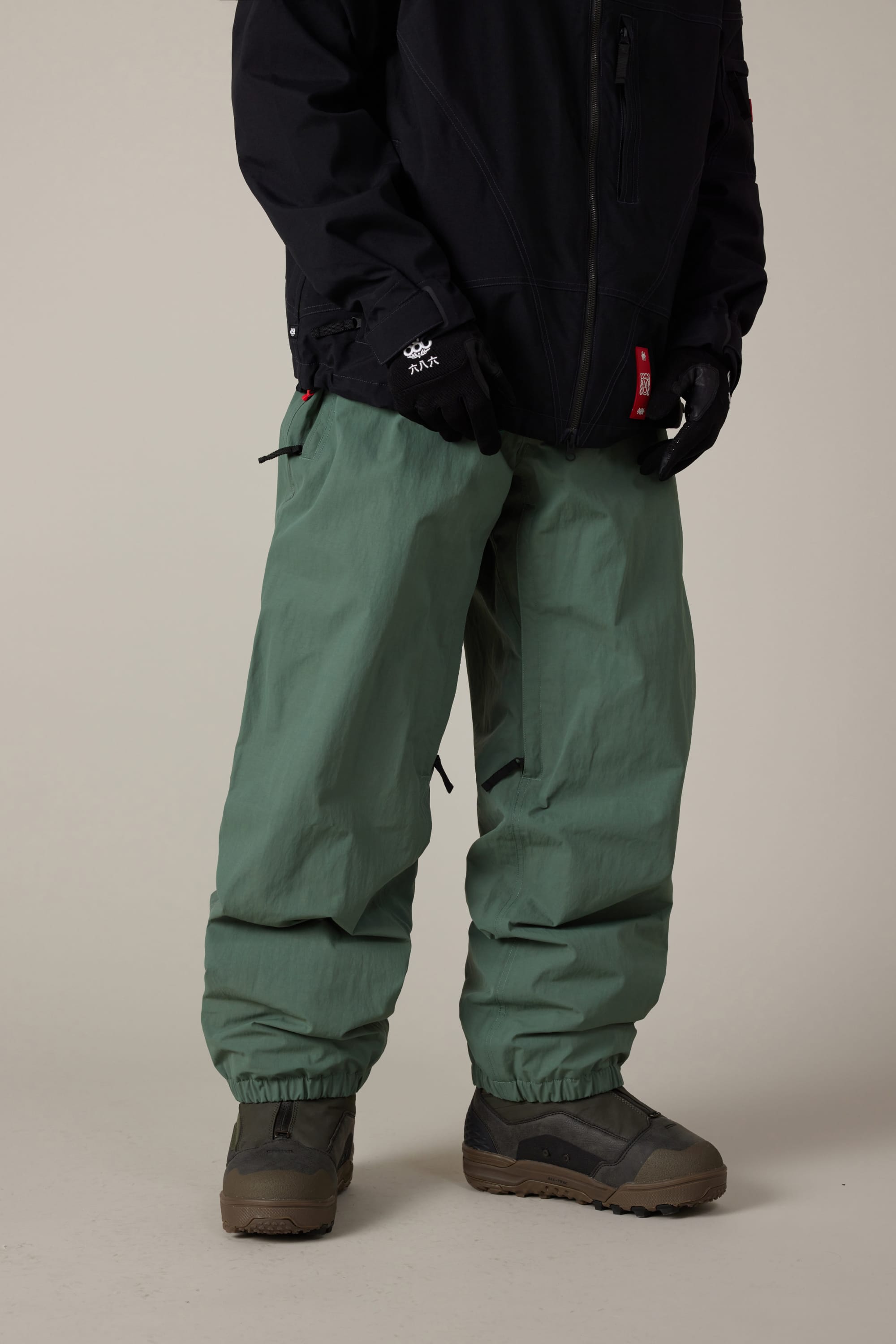 A person wearing the 686 Men’s 686 Dojo® Shell Pant in sage green, paired with a black jacket, black gloves, and dark gray boots stands against a plain background. Their head and upper body are out of the frame.