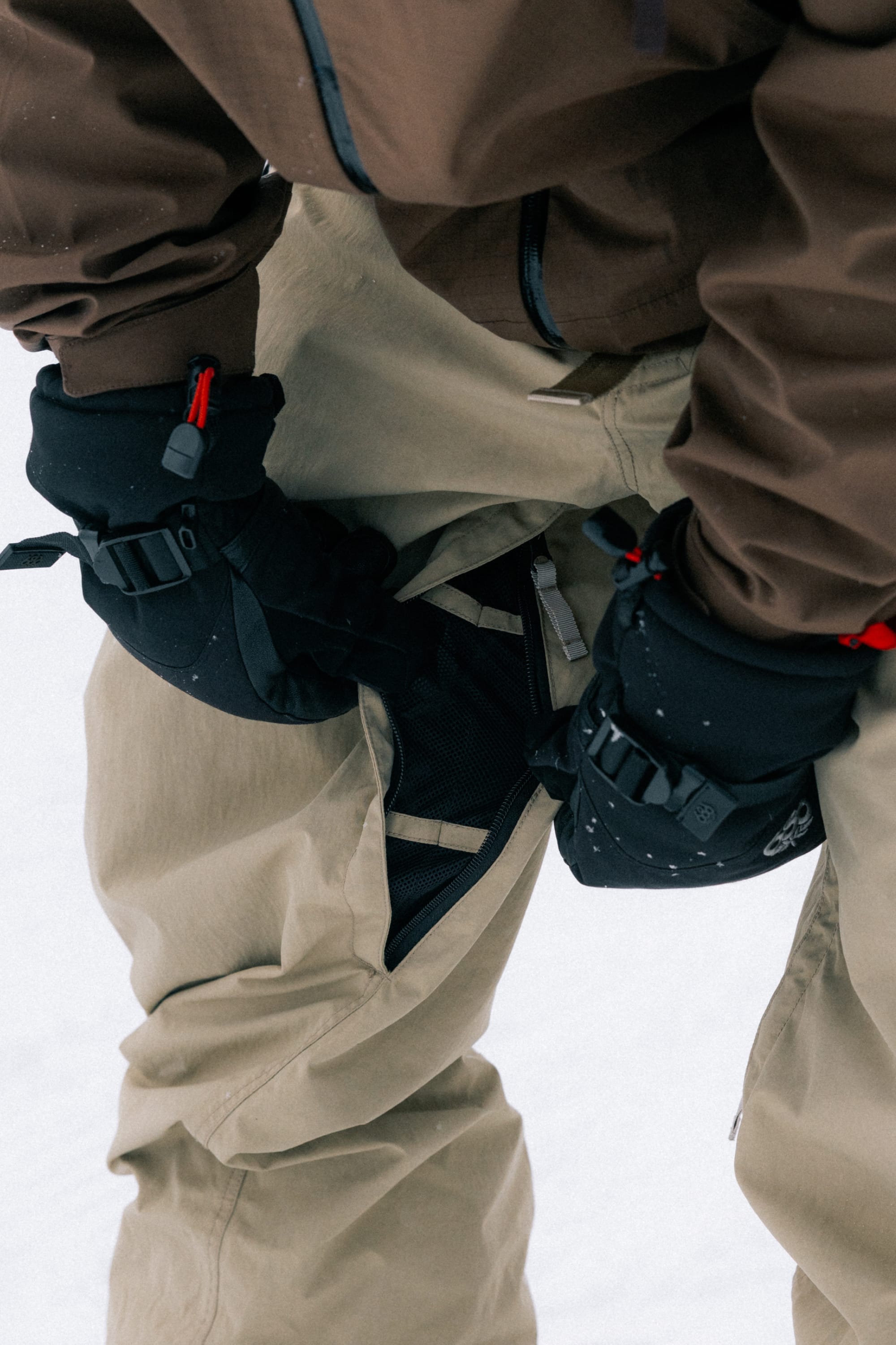 A person wearing 686 Men’s 686 Dojo® Shell Pants in brown and a matching jacket stands in the snow, wearing black winter gloves and opening a zipper on the side of their pants.