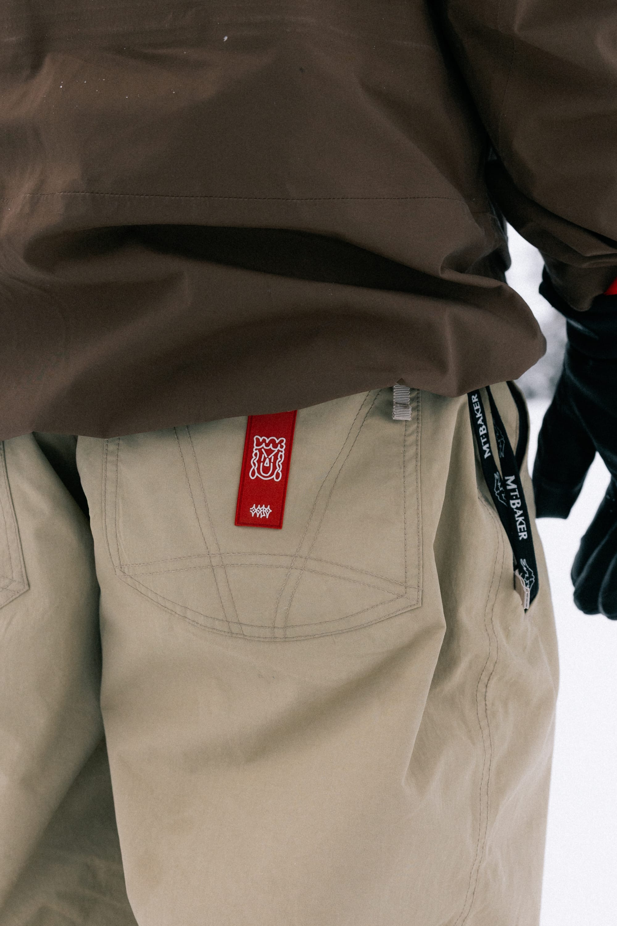 A person wearing a brown jacket and 686 Men’s 686 Dojo® Shell Pant from 686, featuring a red tag on the pocket, stands outdoors. A black lanyard and gloves are visible on the right side.