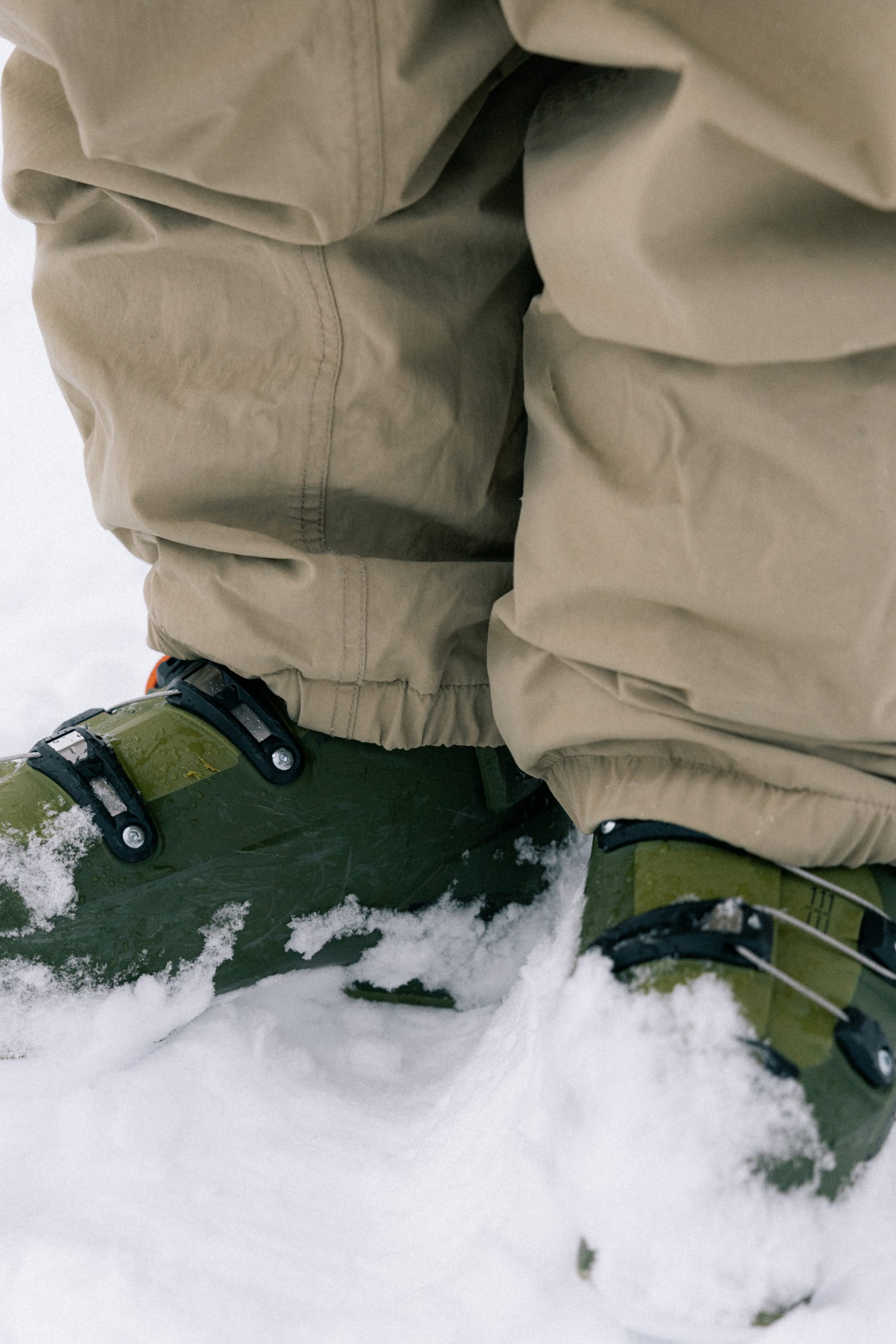 A person wearing the 686 Men’s 686 Dojo® Shell Pant in tan stands in fresh snow, paired with green ski boots.