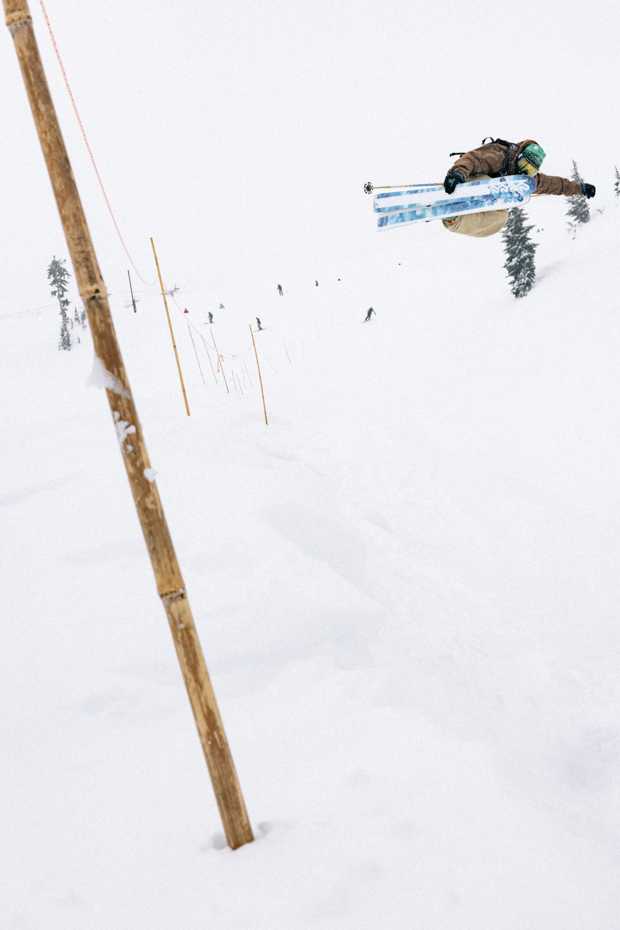 A skier in mid-air performs a trick above snowy slopes, close to a bamboo pole and orange guide rope, wearing the 686 Men’s 686 Dojo® Shell Pant by 686, with trees and distant skiers in the background on a cloudy day.